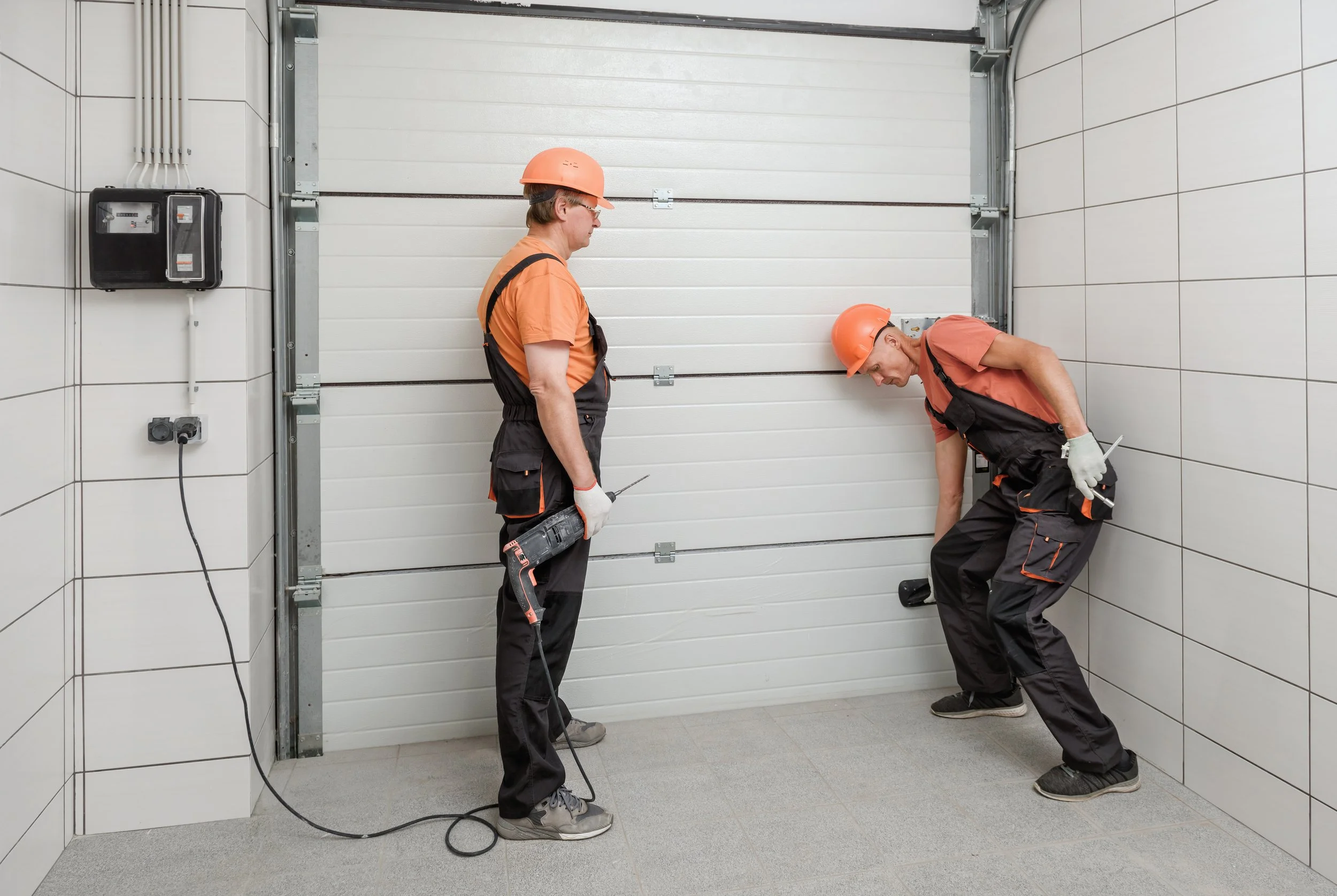 Two workers in orange safety helmets and black work overalls installing or repairing a white garage door inside a small tiled room. One worker stands holding a power drill, while the other is leaning down, possibly fixing or adjusting something.
