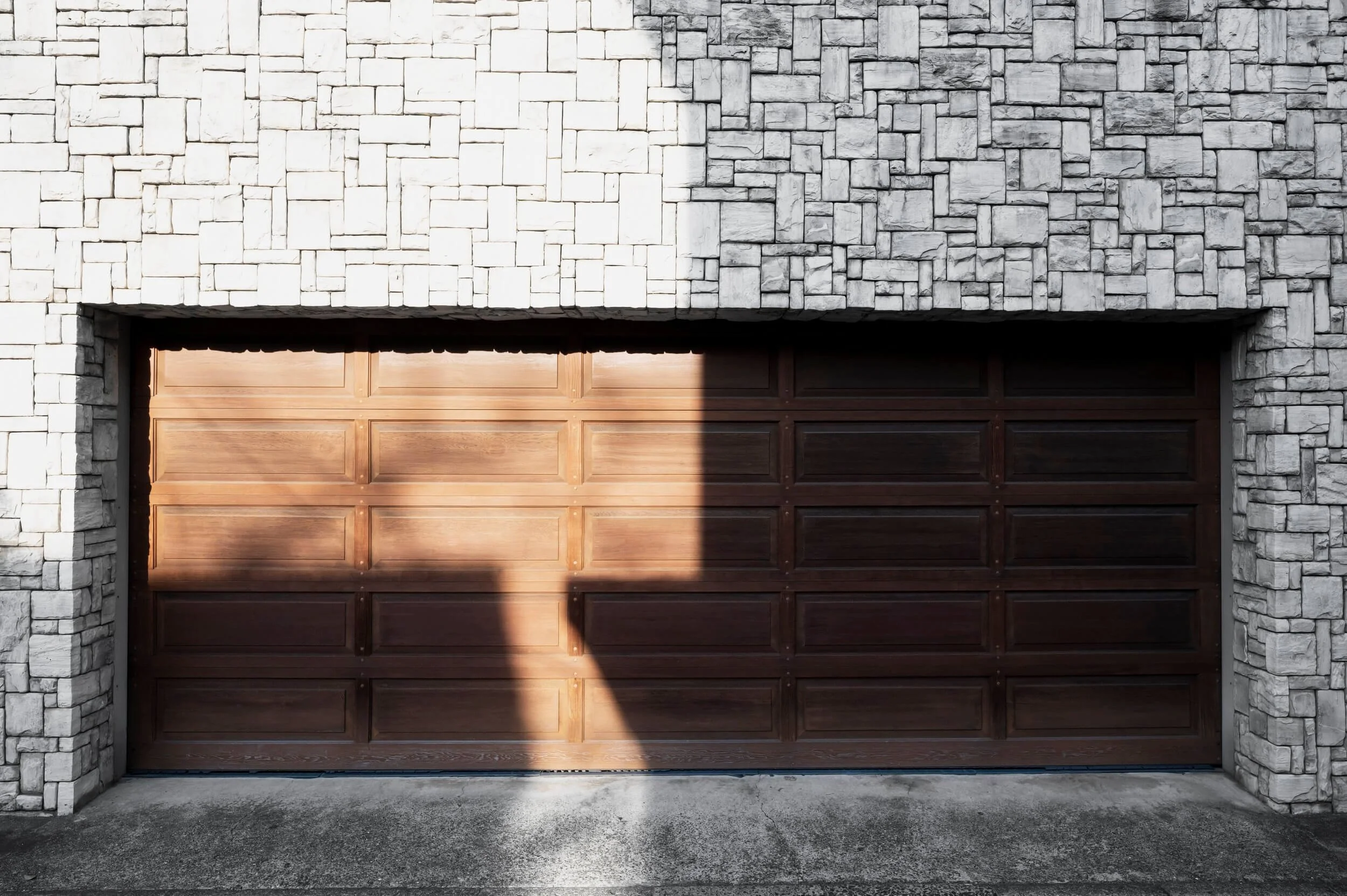 A closed wooden garage door set in a stone brick house facade. The garage door has rectangular panels, with sunlight casting a shadow on the right side.