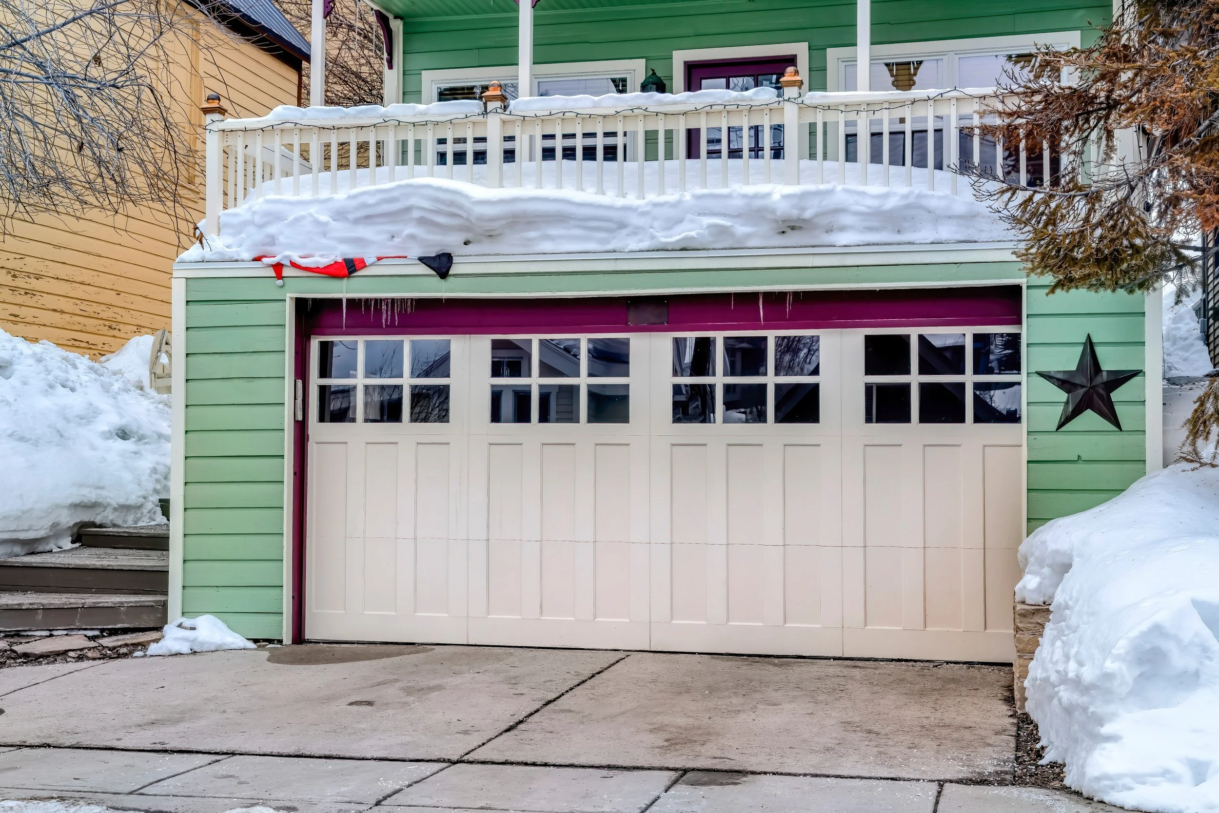 A house with a garage door in front, surrounded by snow, with snow on the roof and a star decoration on the wall to the right of the garage. The house has green siding and a balcony above the garage.