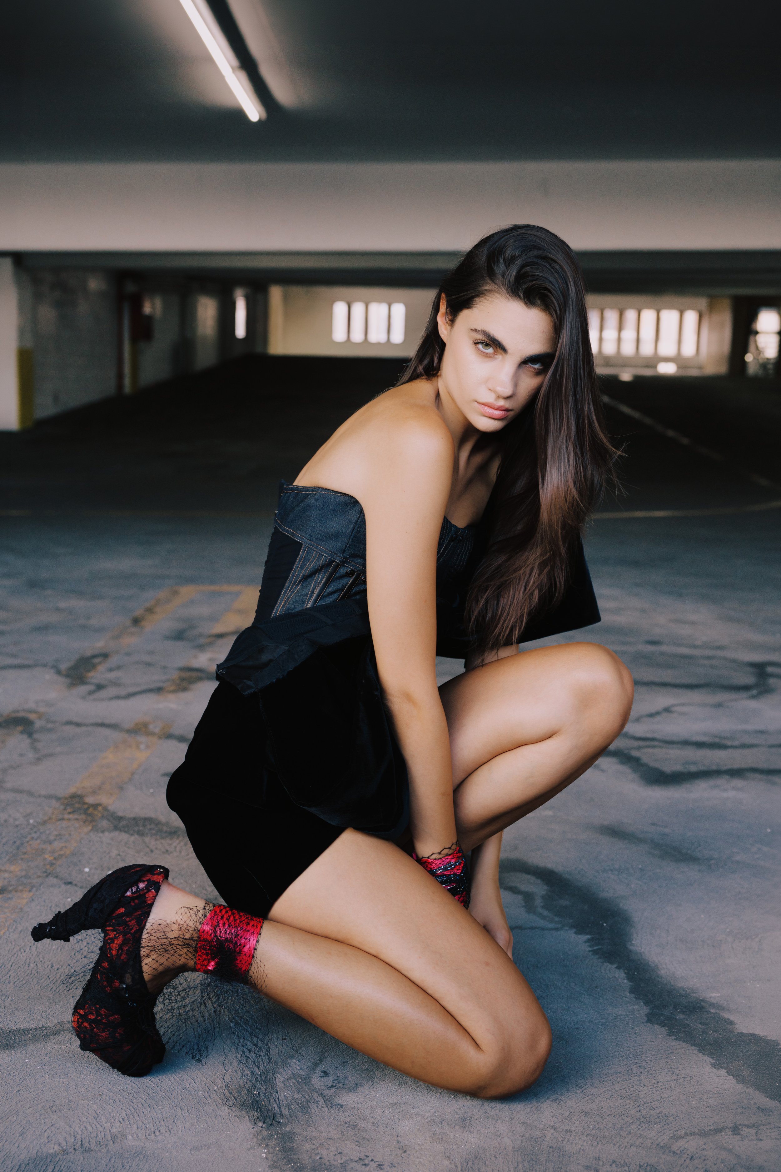 A young woman with long dark hair and intense expression posing in an empty parking garage, wearing a strapless black dress, fishnet socks, and patterned boots.