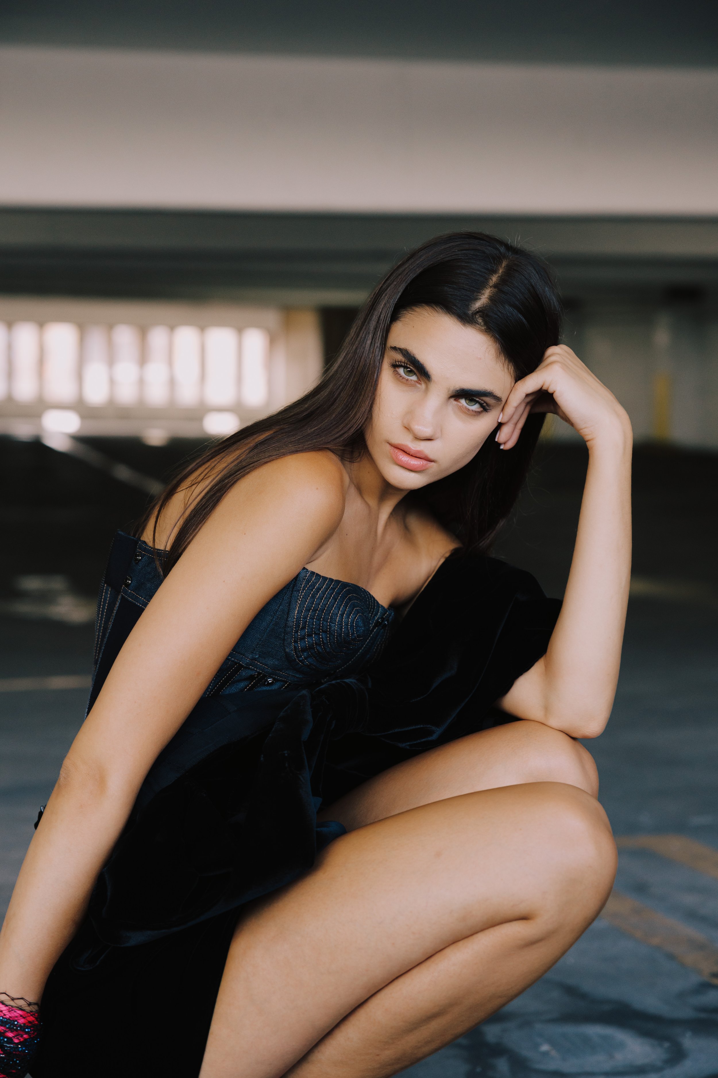 A young woman with long dark hair and striking eyes resembles actor Lana Del Rey, and she is sitting on a parking garage floor with her leg bent and arm resting on her knee, wearing a black sleeveless top.