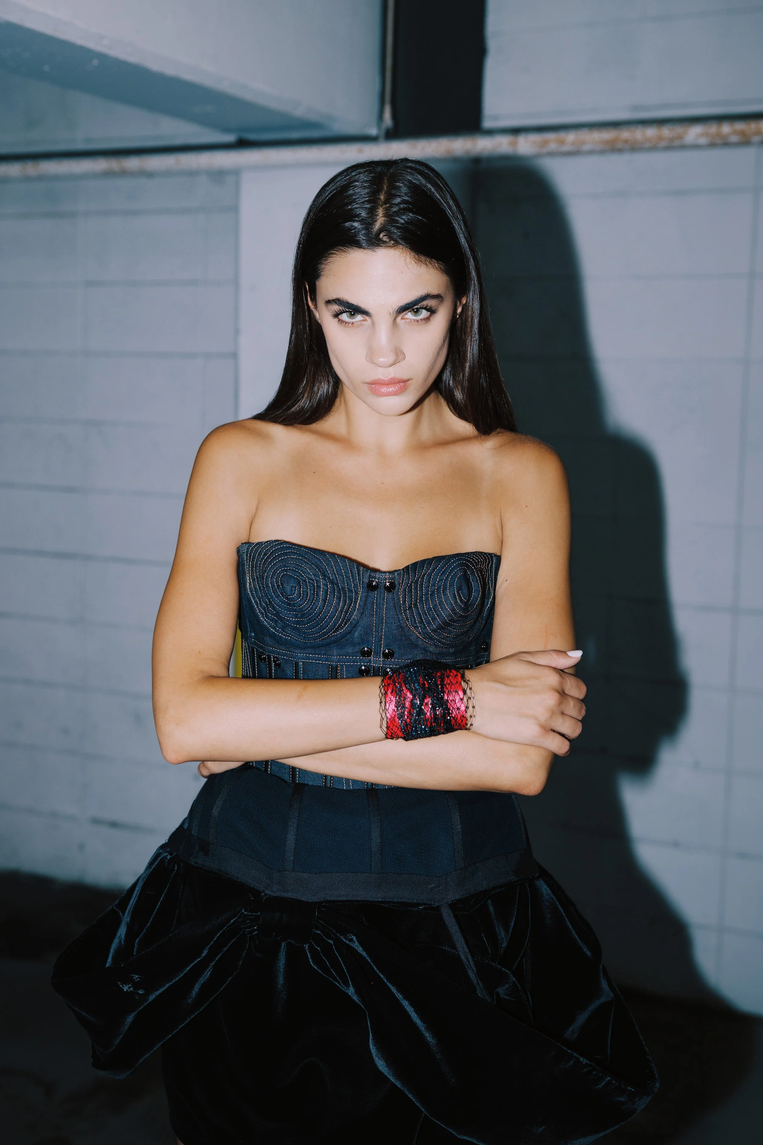 A young woman with dark hair, wearing a strapless black top and a black skirt with a volume effect, stands with her arms crossed. She has a serious expression, light makeup, and is in front of a gray, industrial background.