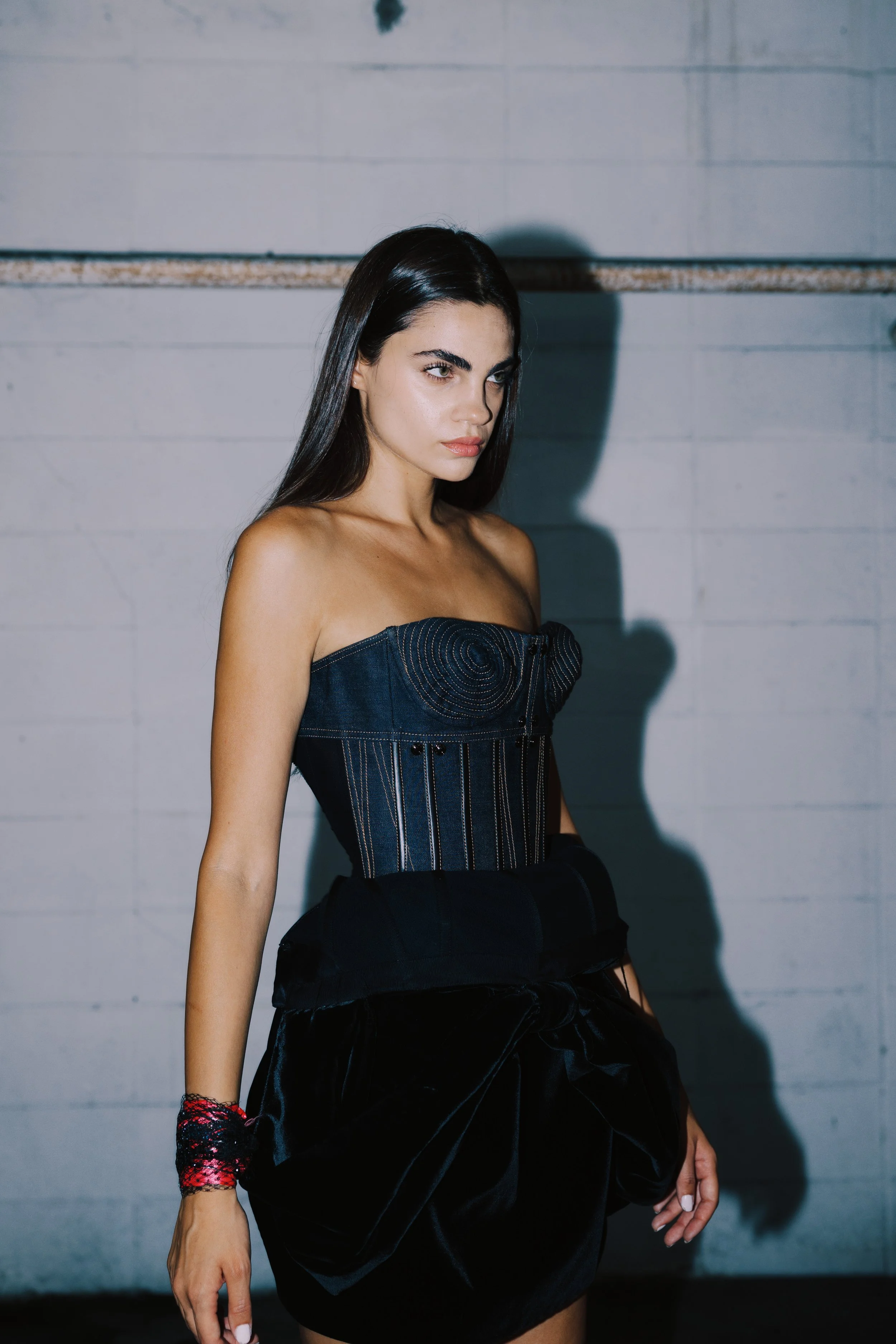 A young woman with long dark hair, wearing a strapless denim top and a black skirt, stands against a white brick wall with a shadow cast behind her. She is gazing slightly to her left with a serious expression.