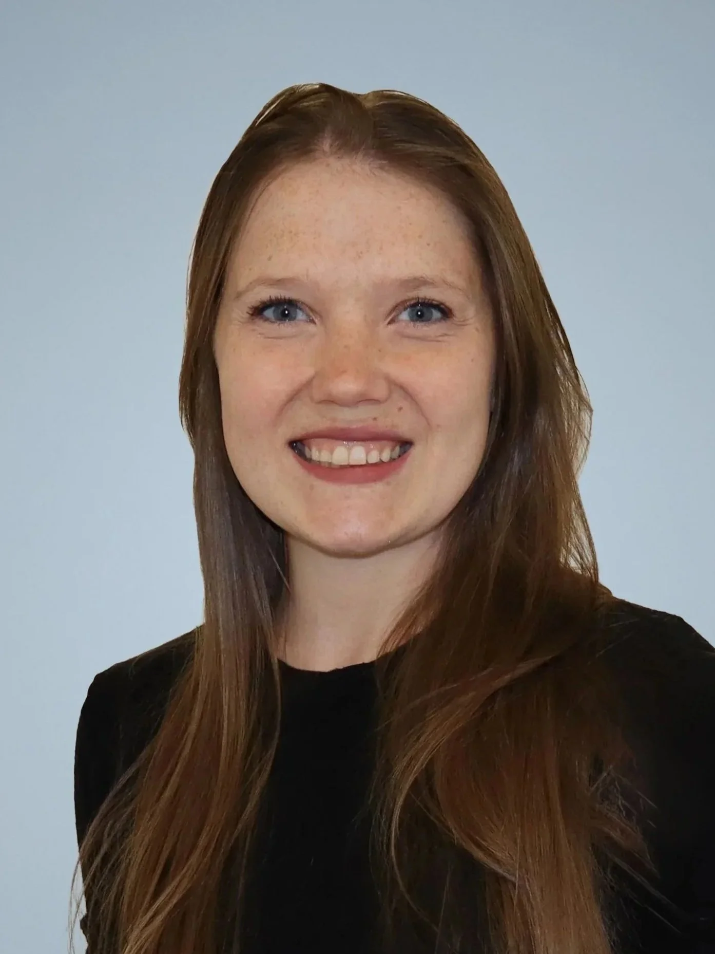 A young woman with long red hair, blue eyes, and freckles smiling against a light gray background.