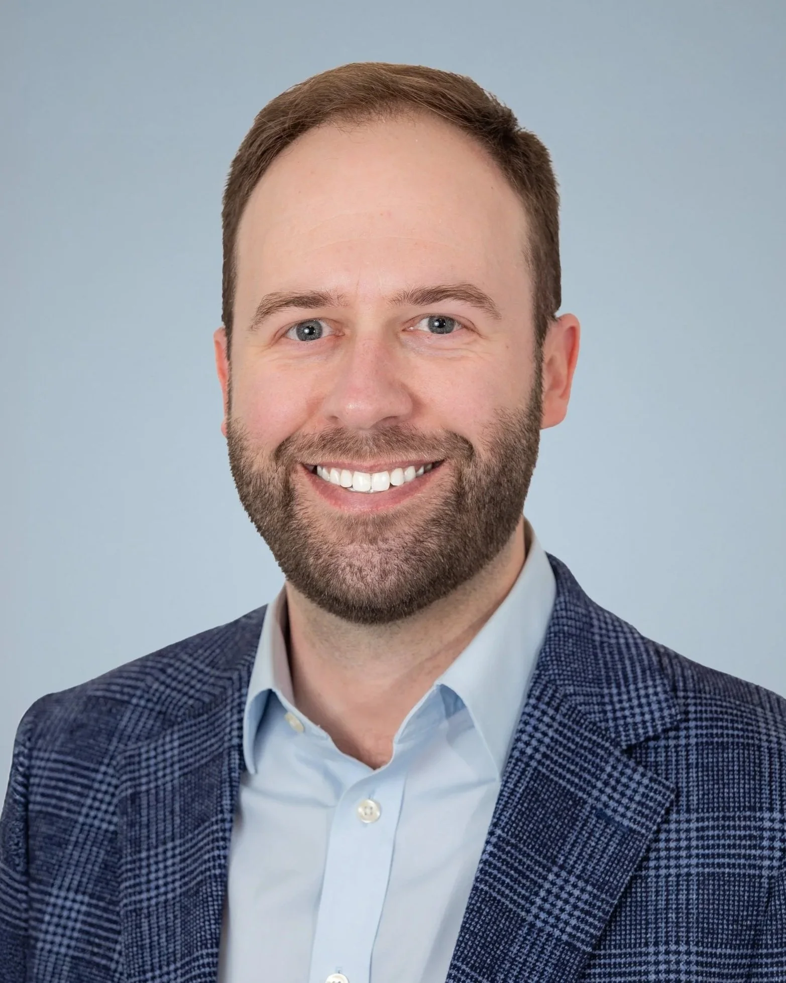 A man with a beard and short brown hair, wearing a blue suit jacket and a light blue collared shirt, smiling in front of a plain, light-colored background.