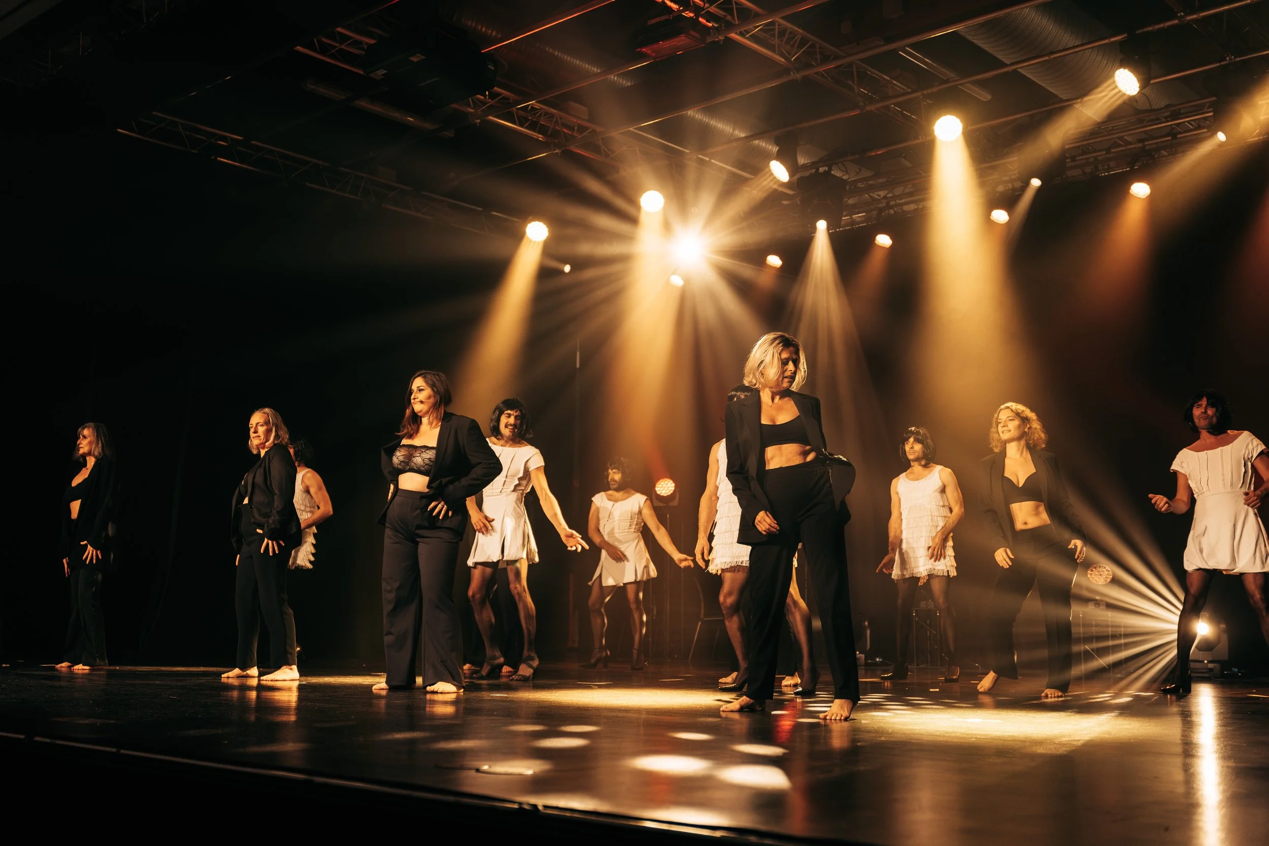 Des femmes exécutent une danse sur scène sous des éclairages chauds, certaines en costumes noirs, d'autres en robes ou tenues blanches, avec un fond sombre.