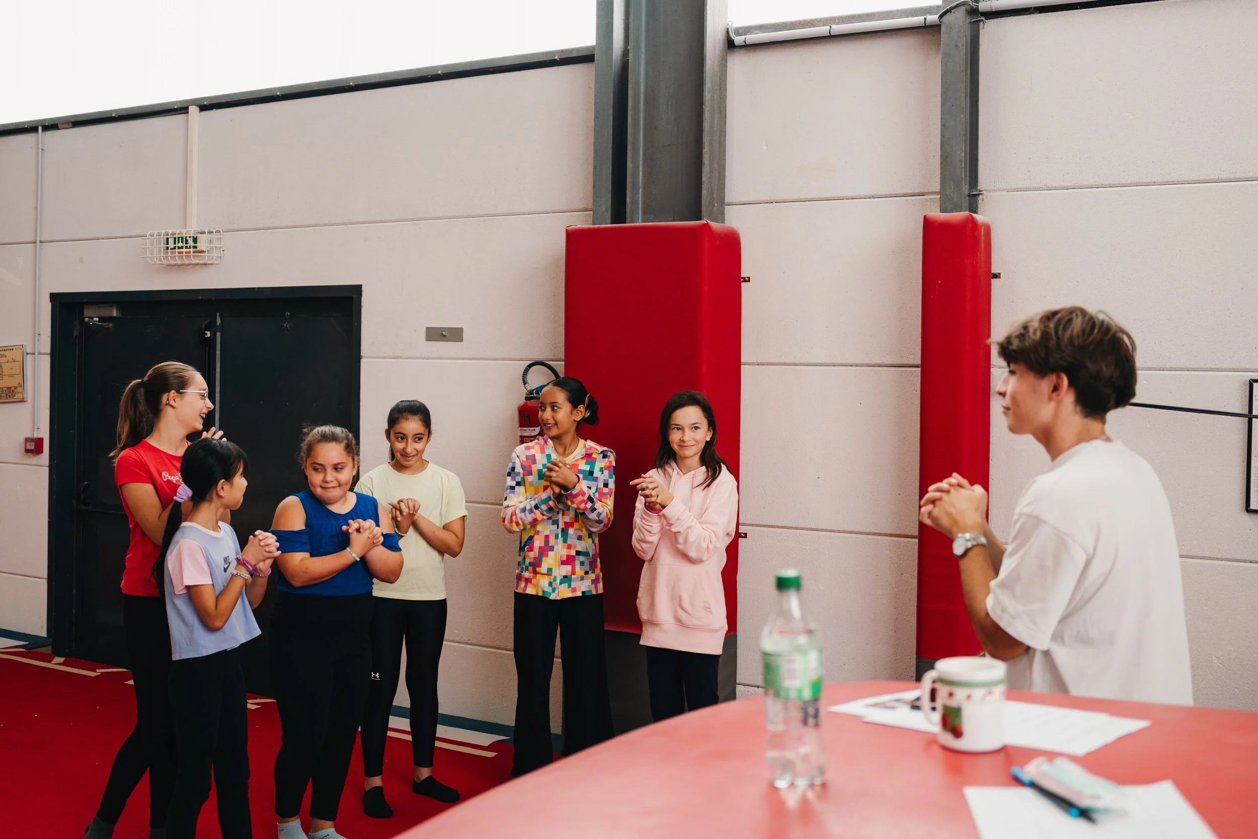 Groupe d'enfants souriants en classe ou en activité, assistant à un enseignant ou animateur qui parle, dans une salle avec murs blanc, tapis rouge et matériel divers.
