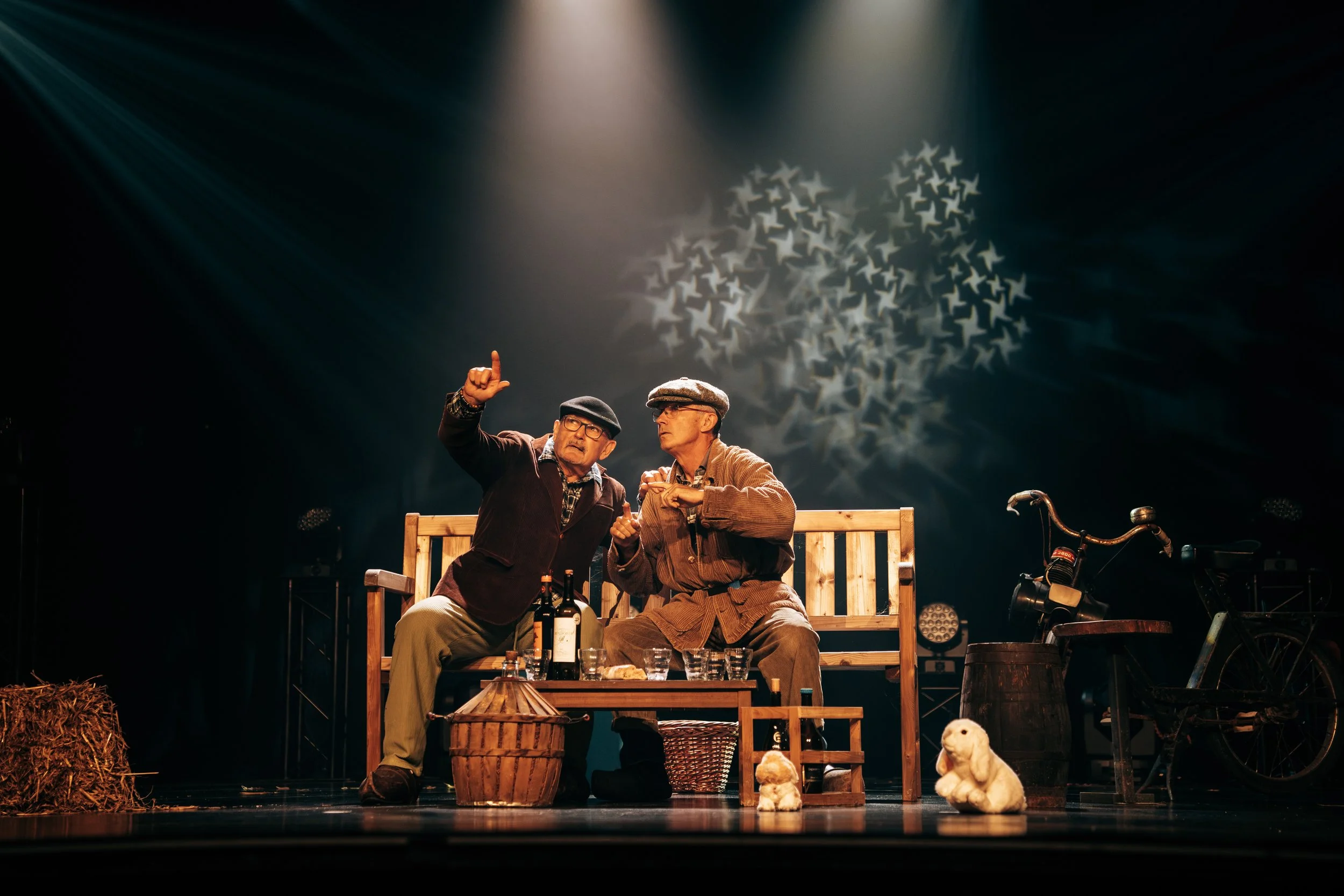 Deux hommes âgés sur scène, assis sur un banc en bois, comme s'ils étaient en train de discuter. L'un d'eux pointe du doigt vers le haut. La scène est décorée avec des bouteilles, des animaux en peluche, un vélo, et un bocal en osier. Un éclairage ta