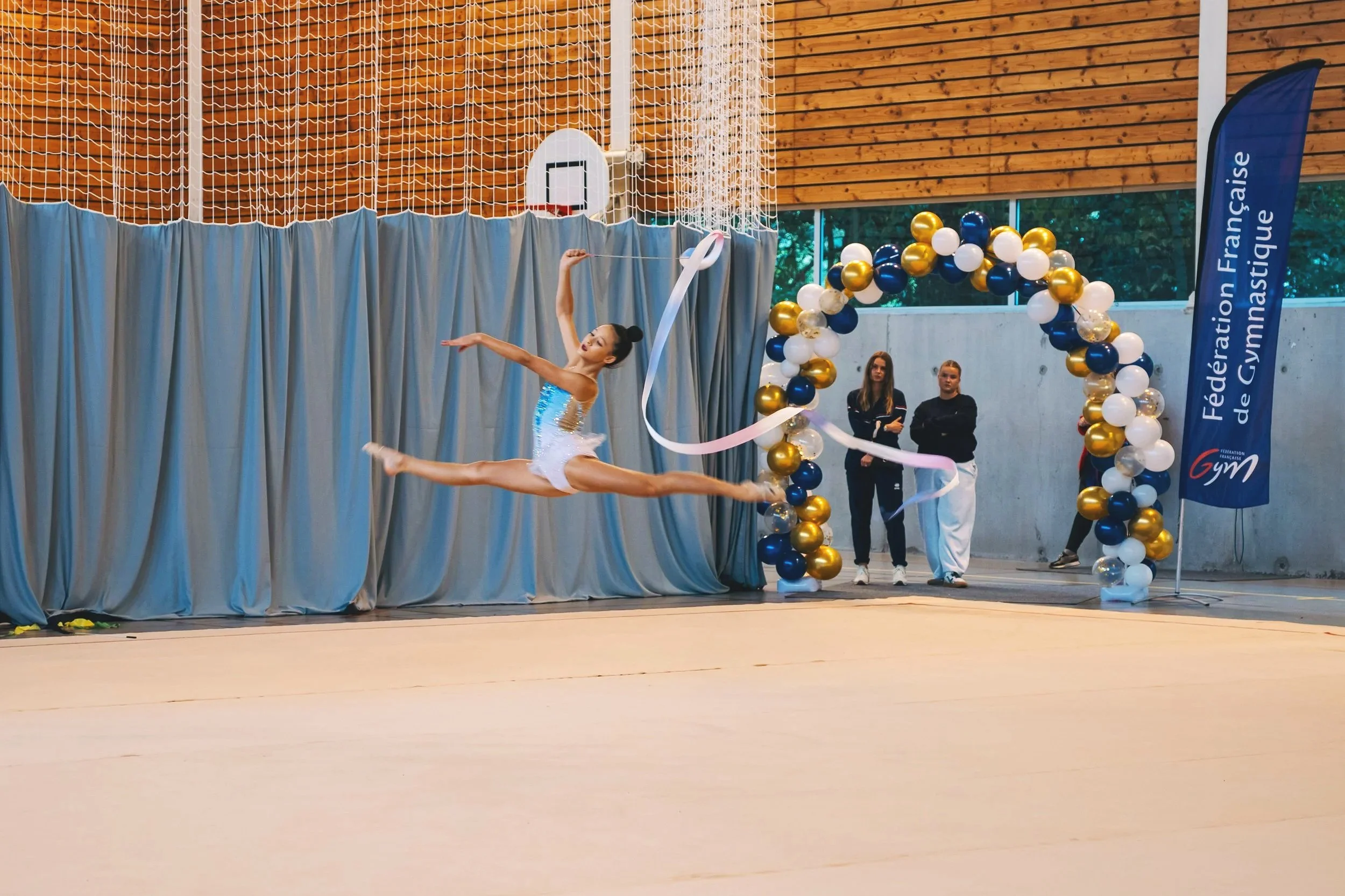 Une gymnaste effectue une figure en saut en plein air lors d'une compétition, avec un décor de ballons bleus, blancs et or et un drapeau de la Fédération Française de Gymnastique.