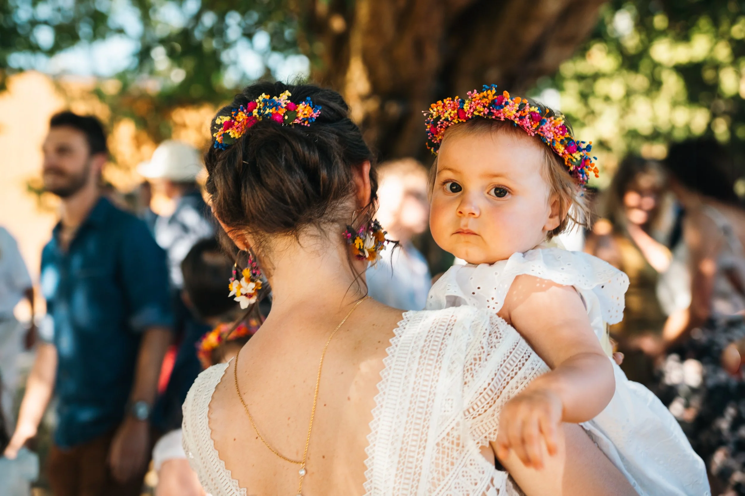 Une femme portant une robe blanche et des accessoires floraux, tenant un jeune enfant portant une robe blanche et un floral de fête sur la tête, lors d'une célébration en plein air sous un arbre.