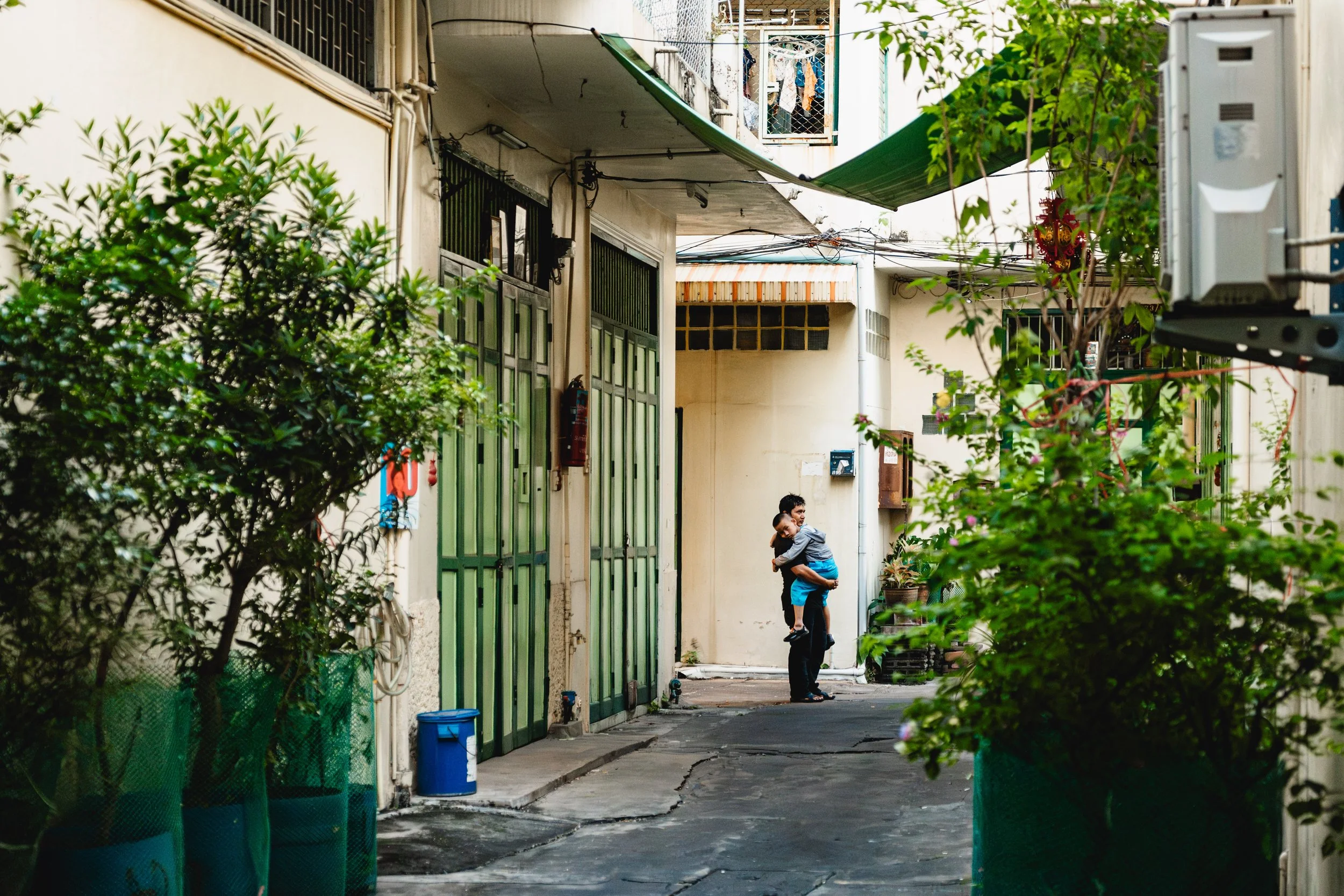 Deux enfants s'embrassent dans une ruelle urbaine, entourée de plantes en pots, avec des bâtiments en arrière-plan.