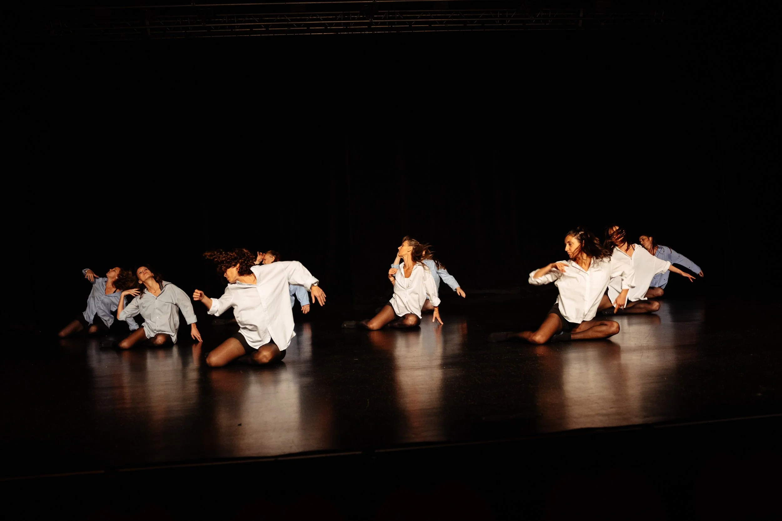 Dix danseuses sur scène, habillées en chemises blanches et bleues, en mouvement dans une performance de danse contemporaine, avec un fond noir.