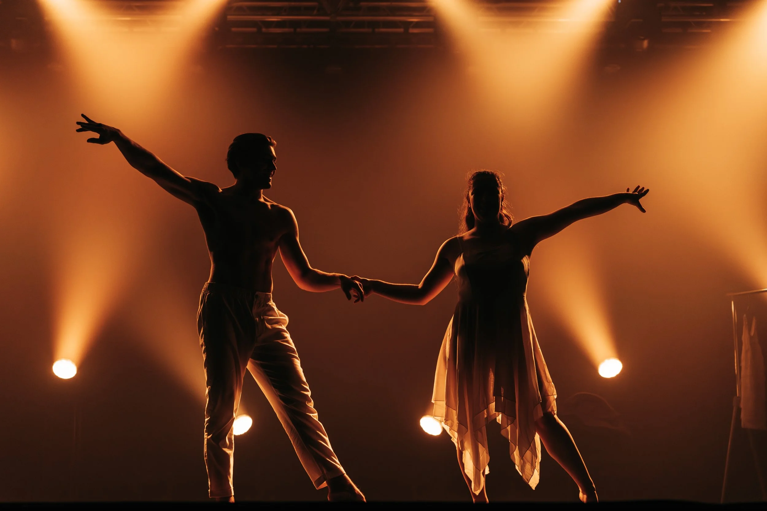 Deux danseurs en silhouette sur scène éclairés par des projecteurs orange, l'homme est chauve et porte un pantalon léger, la femme porte une robe fluide, ils se tiennent par la main dans une pose de danse expressive.