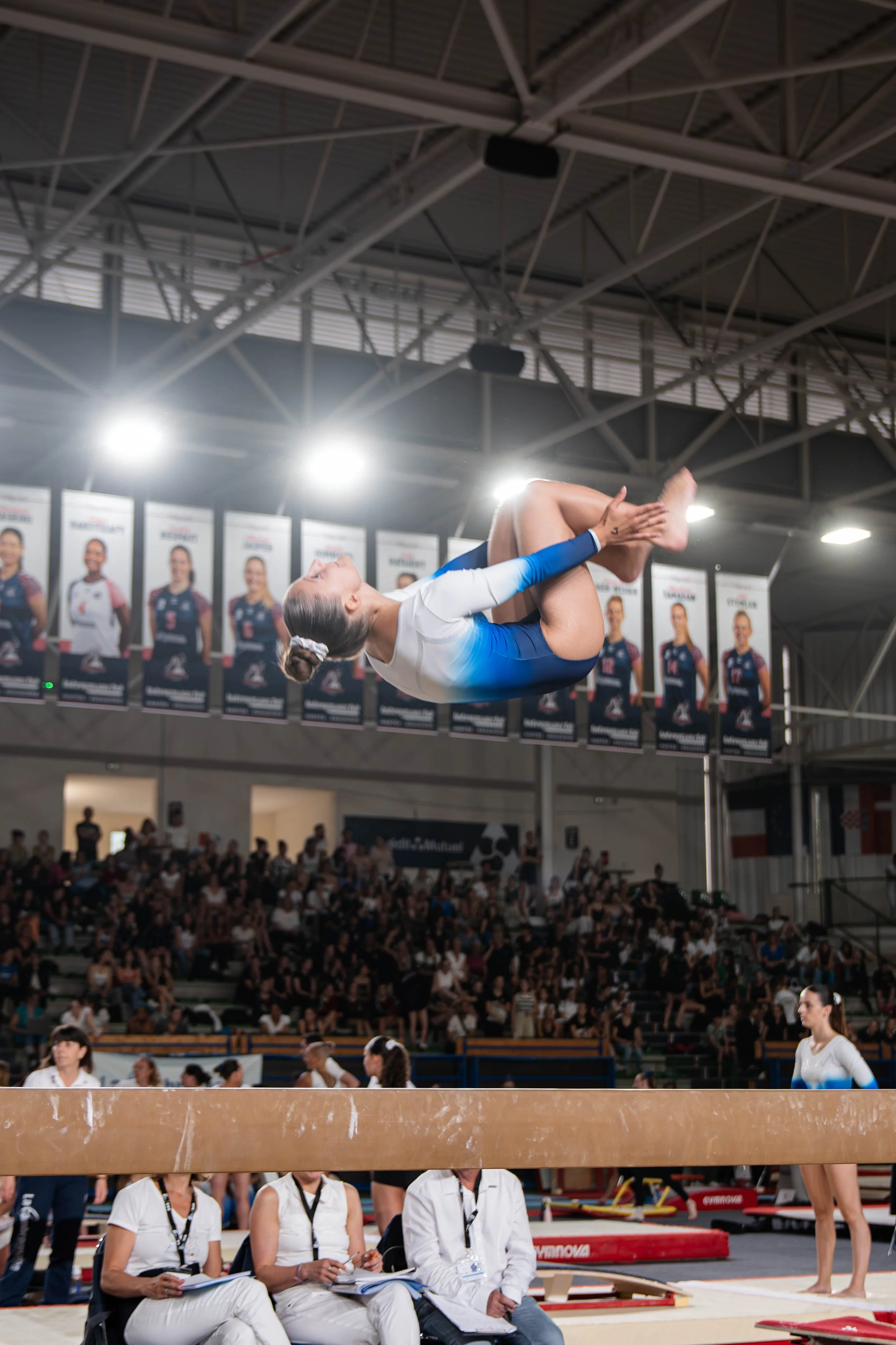 Une gymnaste effectuant un saut en plein air à l'intérieur d'une salle d'entraînement, avec un public et des juges en arrière-plan.