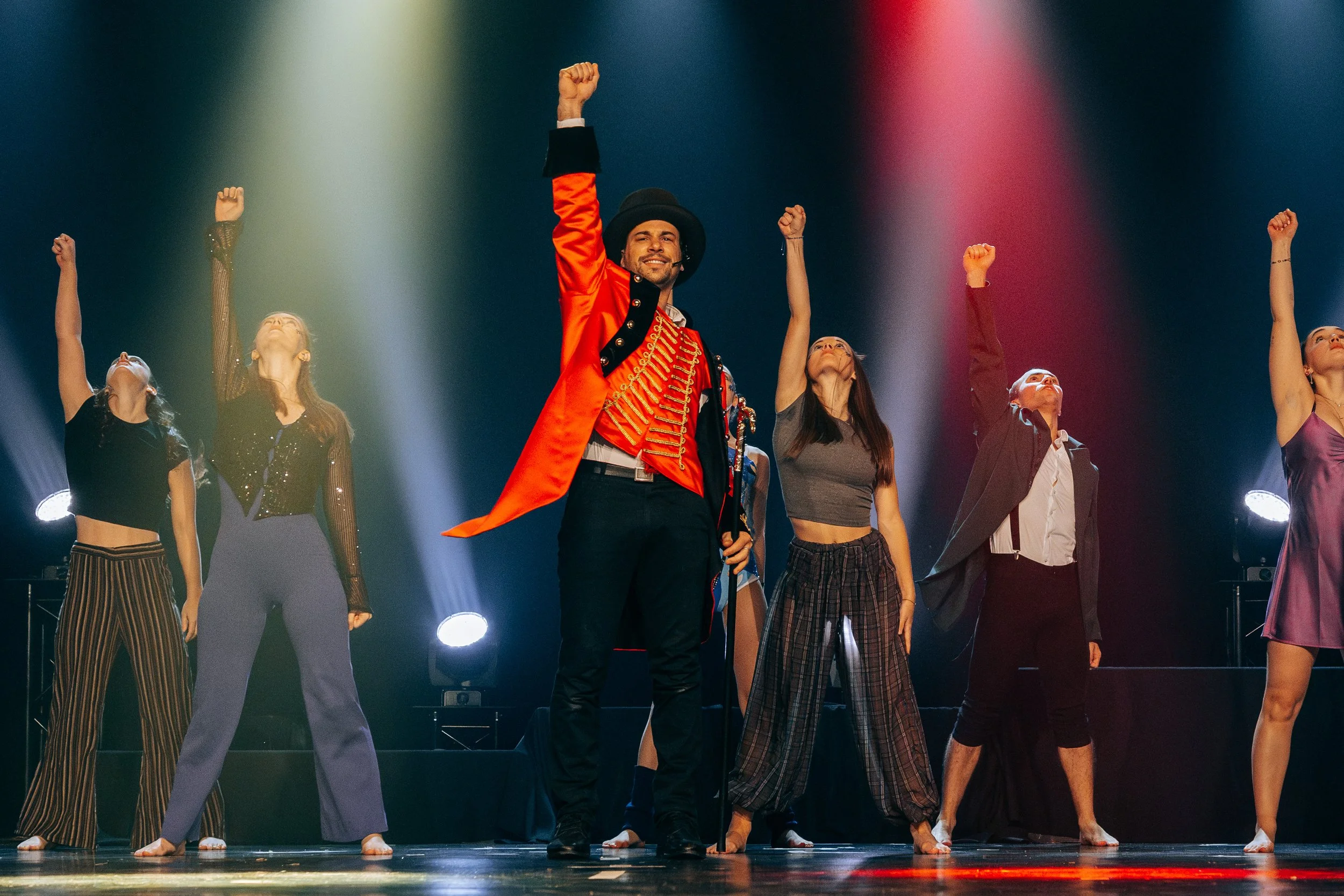 Un performers en costume clown sur scène, entouré de danseuses, avec un éclairage de scène coloré.