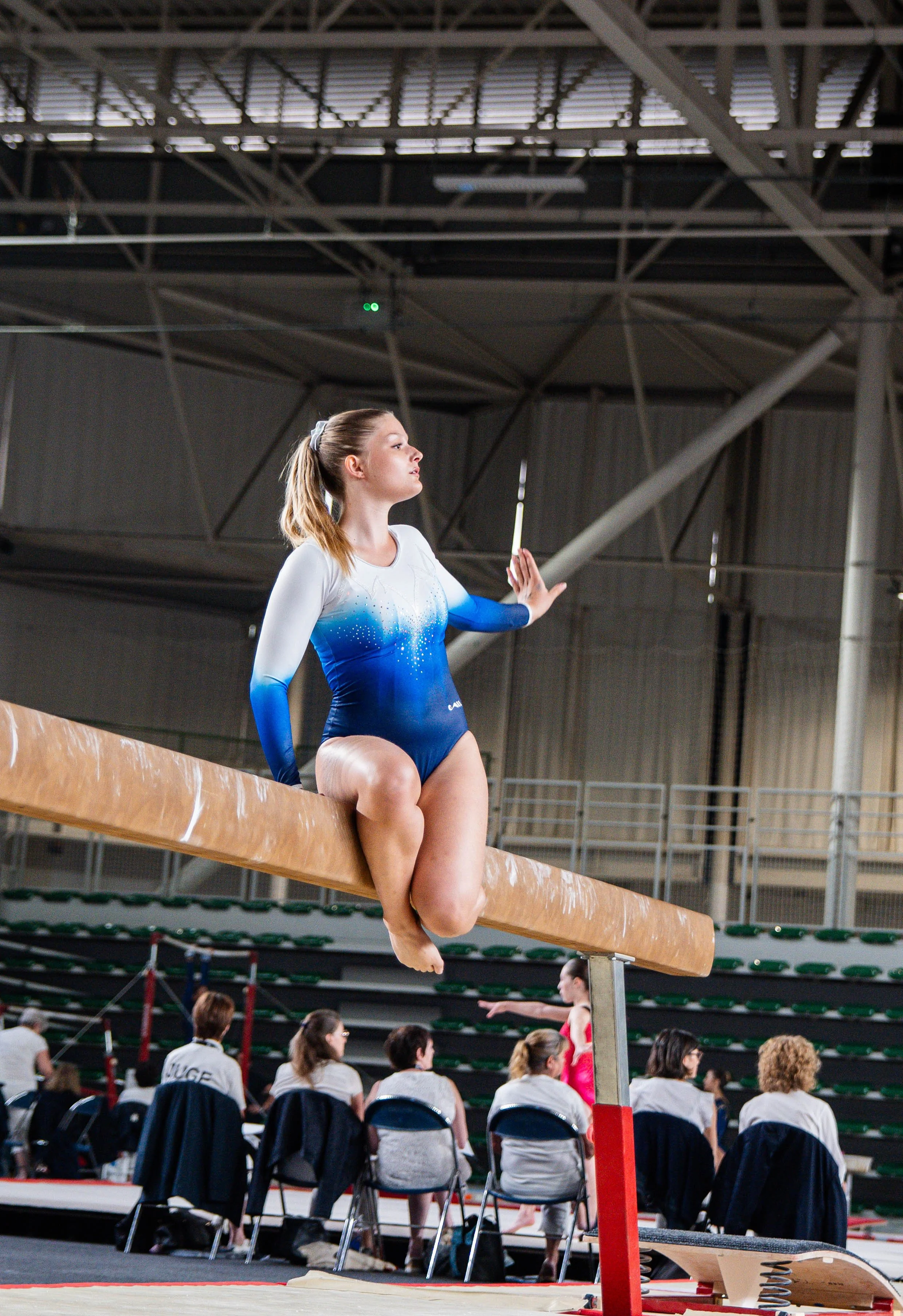 Une jeune gymnaste en uniforme bleu et blanc, assise sur une poutre de gymnastique, en train de se concentrer avant une performance dans une salle d'entraînement ou de compétition.
