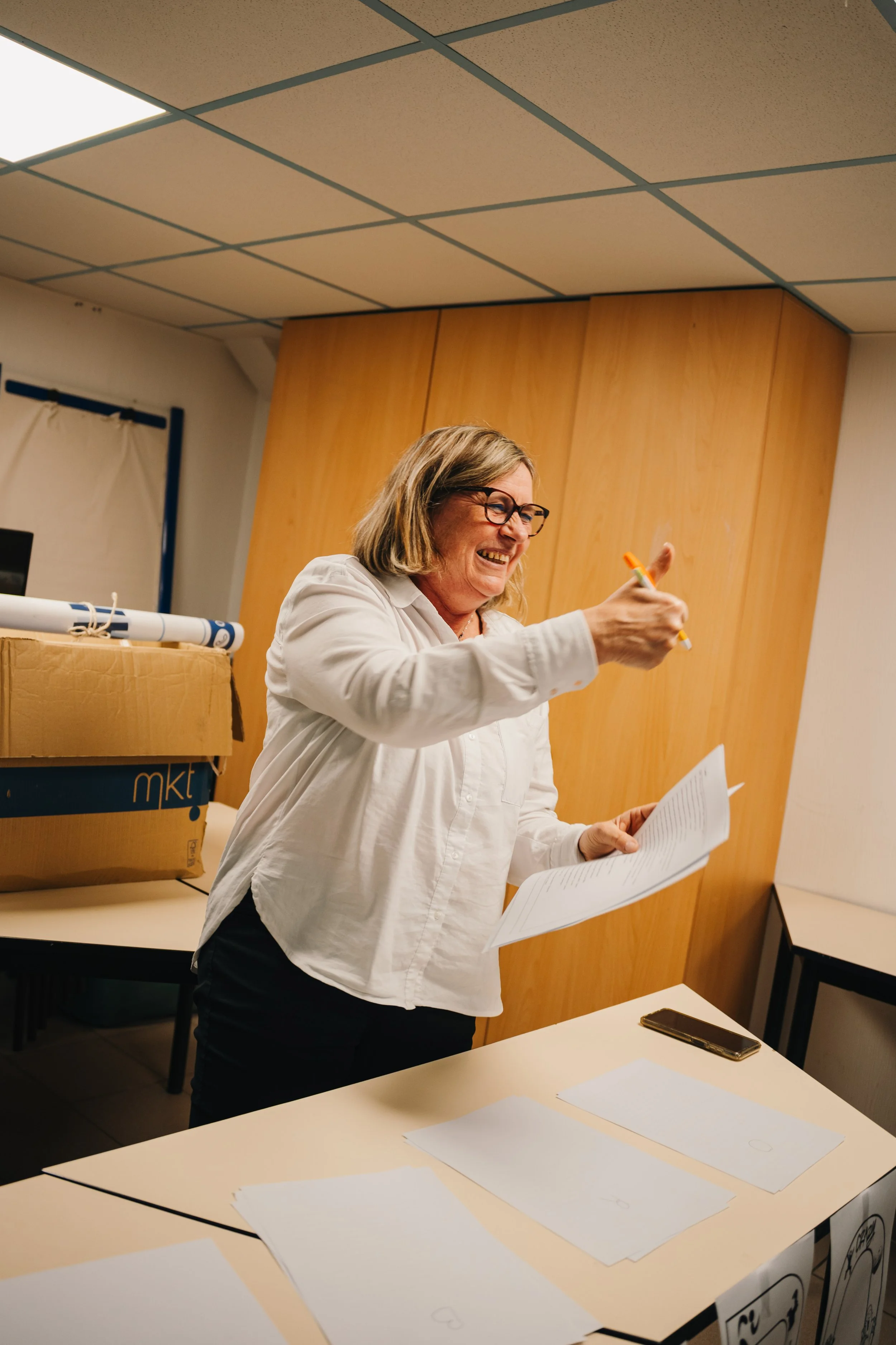 Une femme souriante, portant des lunettes, tient un stylo orange et un document, dans une salle de réunion ou de classe.