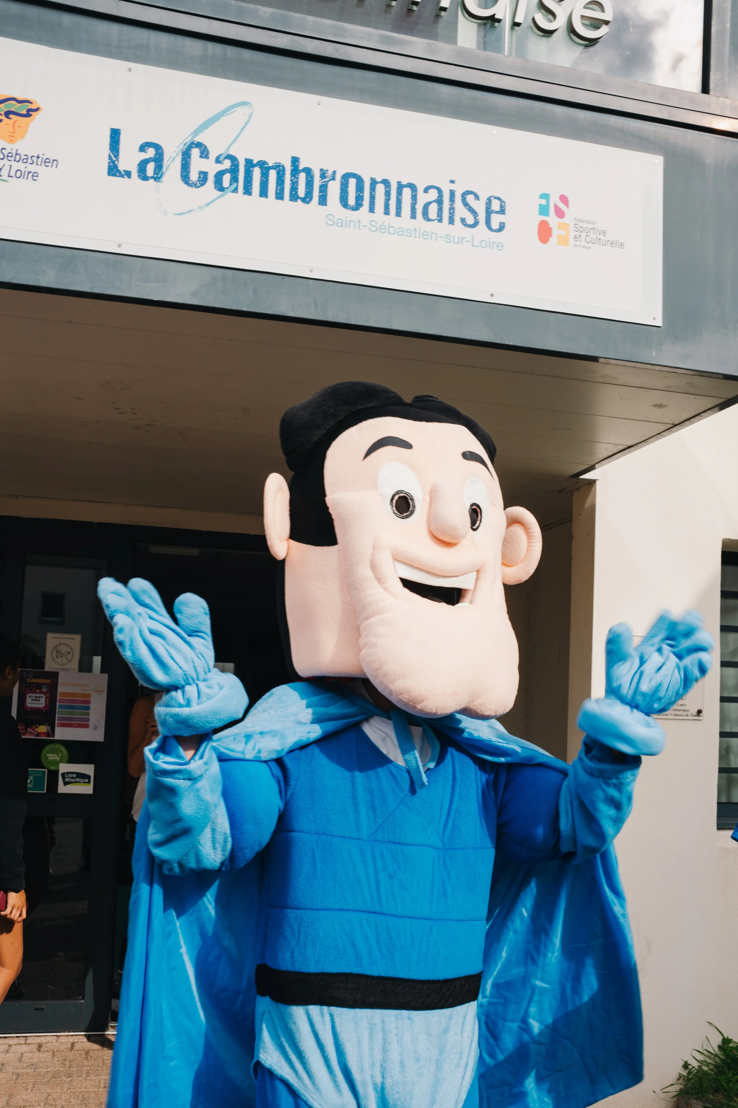 Mascotte en costume de personnage cartoon avec un costume bleu et cape, applaudissant devant le bâtiment de La Chambronnaise à Saint-Sébastien-sur-Loire.