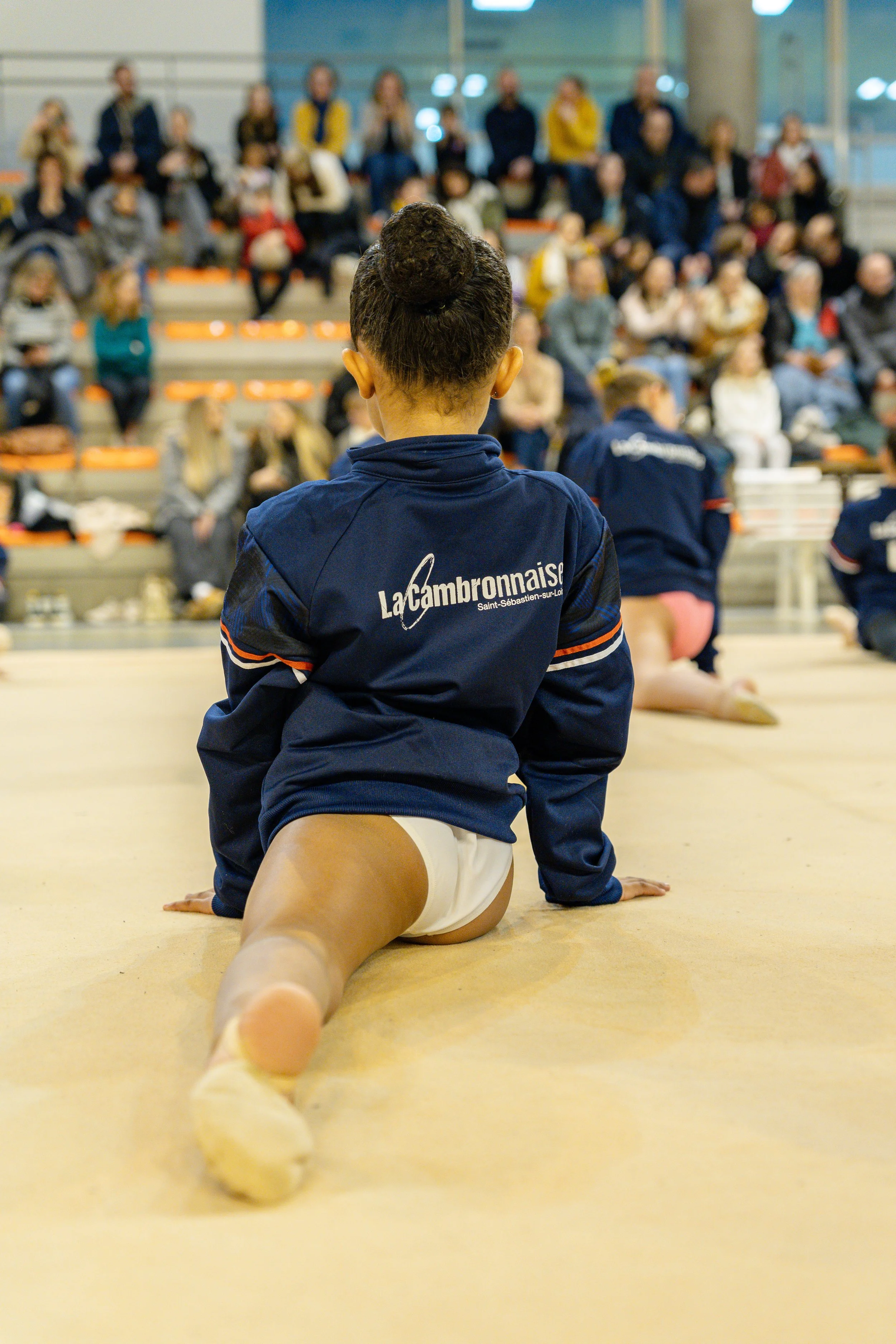 Fille en tenue de gymnastique assise sur le sol d'un gymnase, vue de derrière, avec un groupe de spectateurs assis en hauteur en arrière-plan.