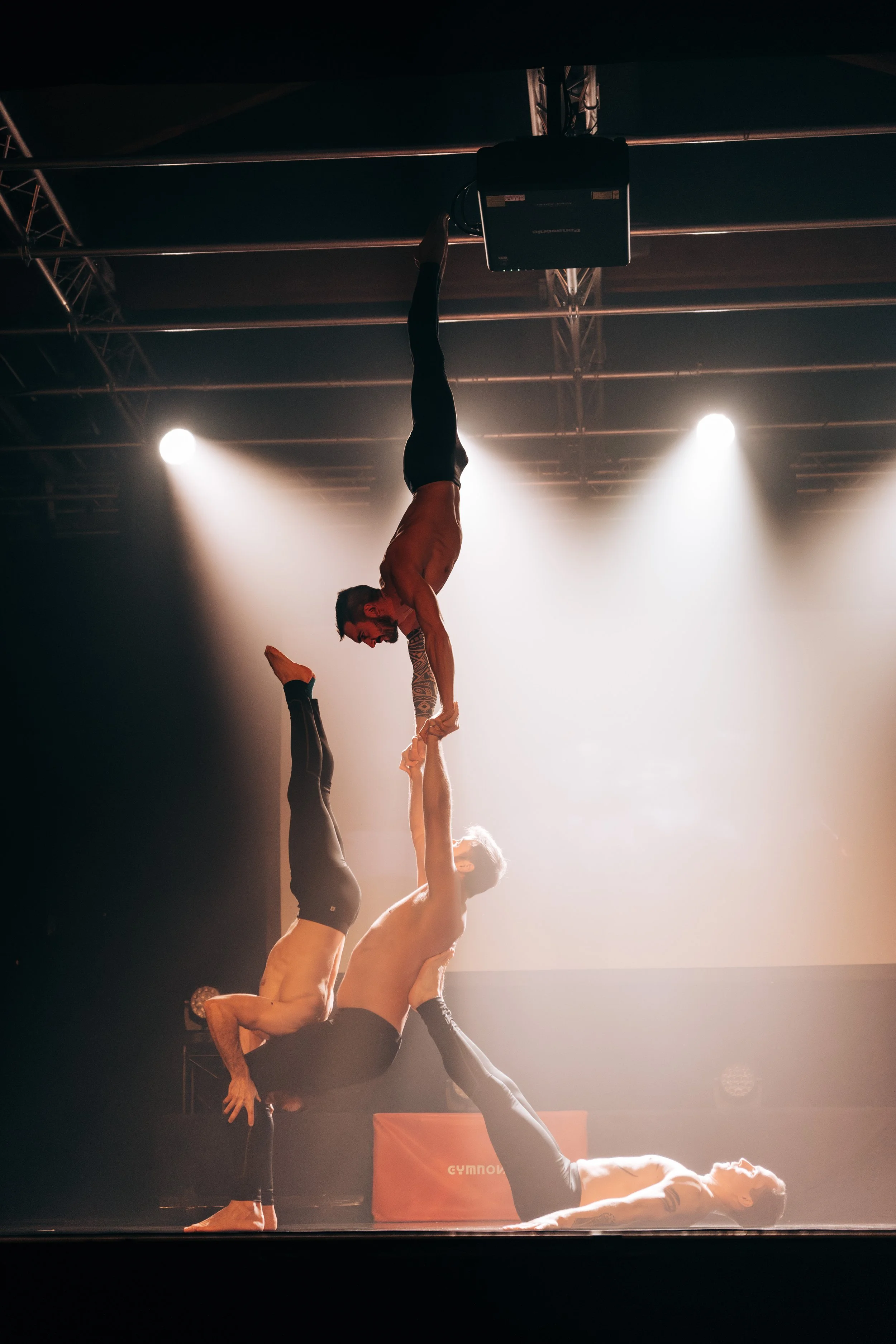 Trois hommes pratiquant le yoga acrobatique sur scène avec un éclairage dramatique.