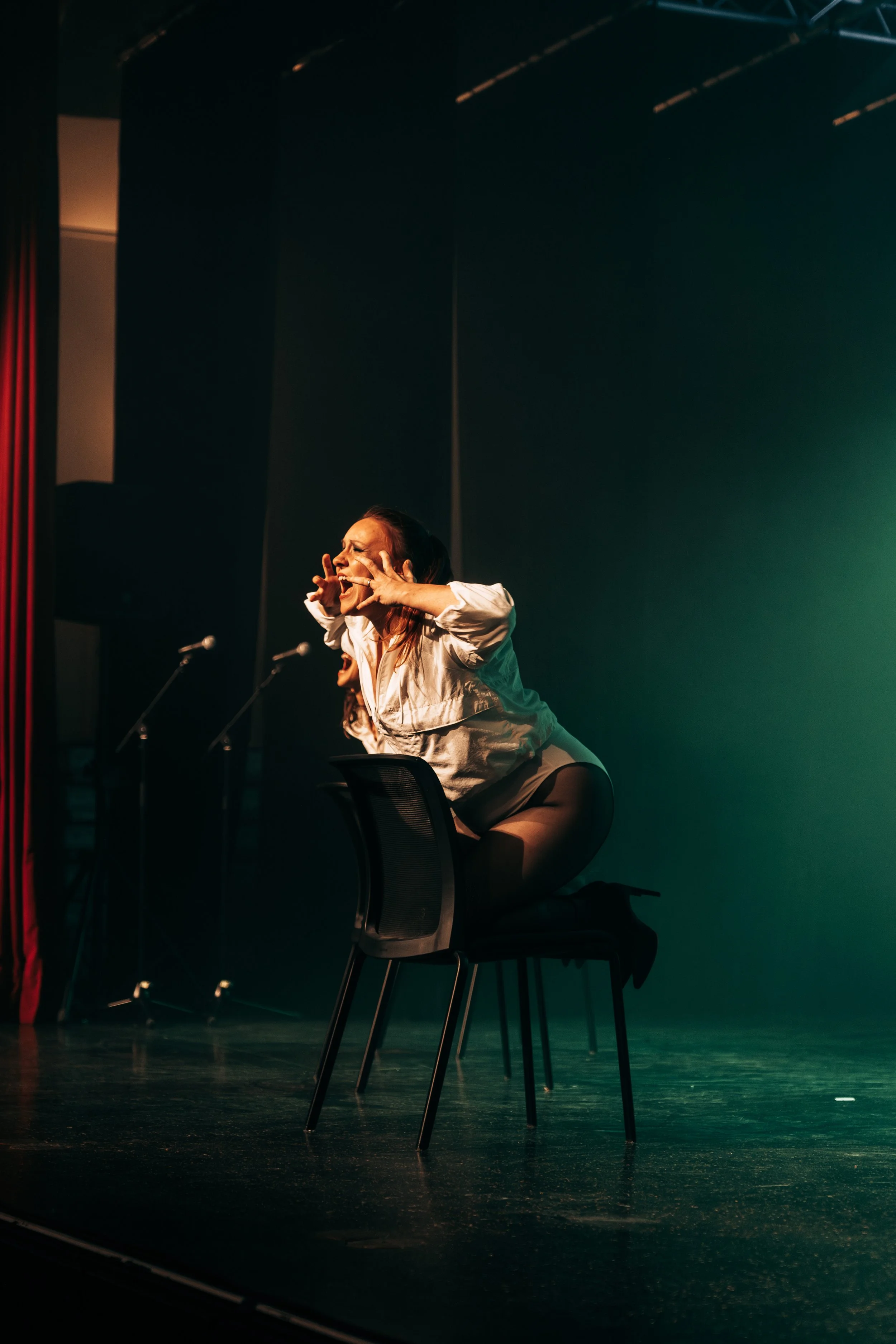 Une femme en talons hauts et jupe noire, assise sur une chaise, fait une expression intense sur scène, avec un fond sombre et une lumière verte.