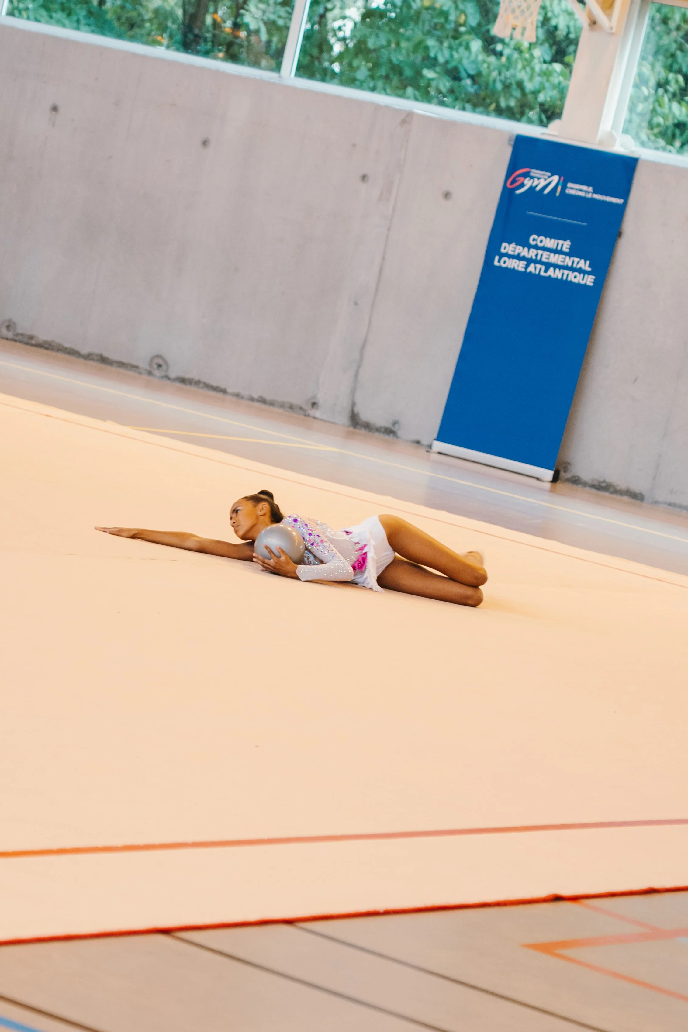 Une jeune gymnaste en tenue de compétition posant sur un tapis de gymnastique avec un ballon, dans une salle de gym avec une bannière bleue en arrière-plan.
