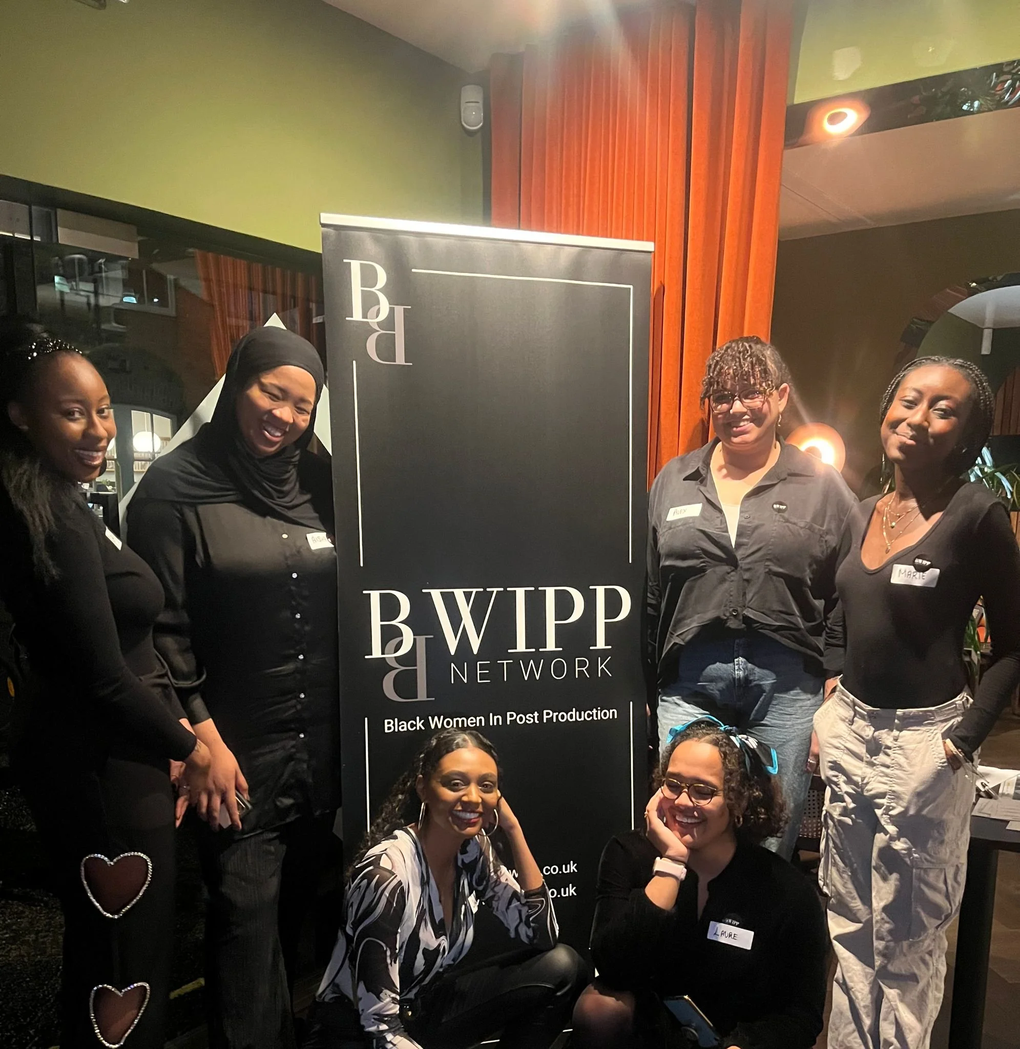 Group of seven women at a Black Women in Post Production event, with some standing and two sitting in front, all smiling, in front of a black banner with white text for BWIPP Network.