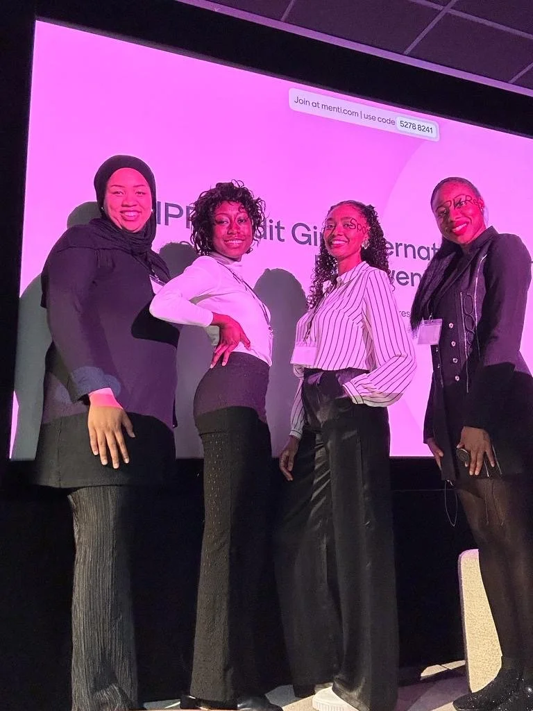 Four women standing on a stage in front of a pink screen, smiling and posing for a photo. The screen has text that says "Join at menti.com | use code 5278 8241" and some partially visible text.