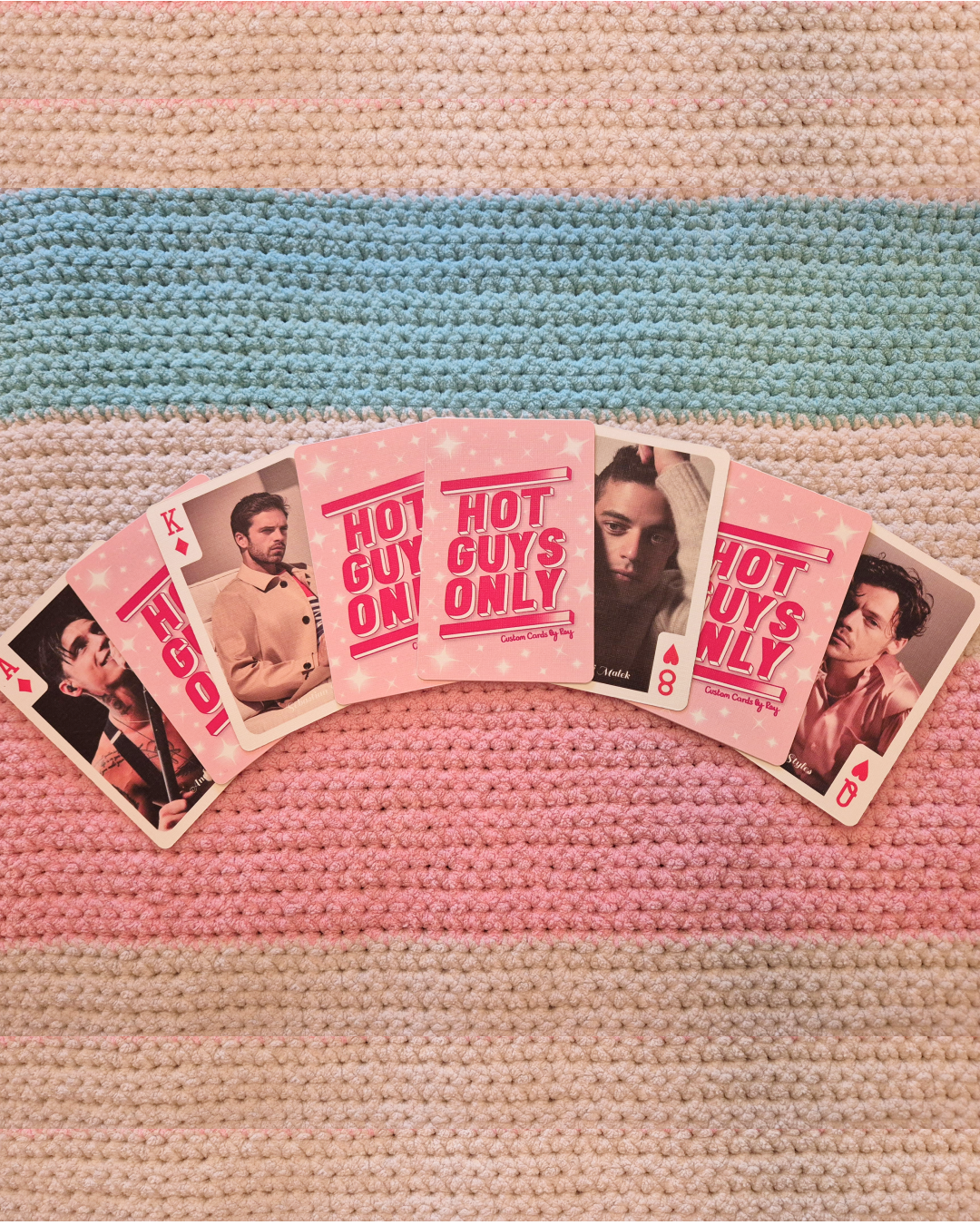 A set of playing cards arranged in a fan on a multicolored, striped, textured blanket. The cards include the king of diamonds, the ace of hearts, and the eight of hearts, with center cards displaying pink text 'HOT GUYS ONLY'.