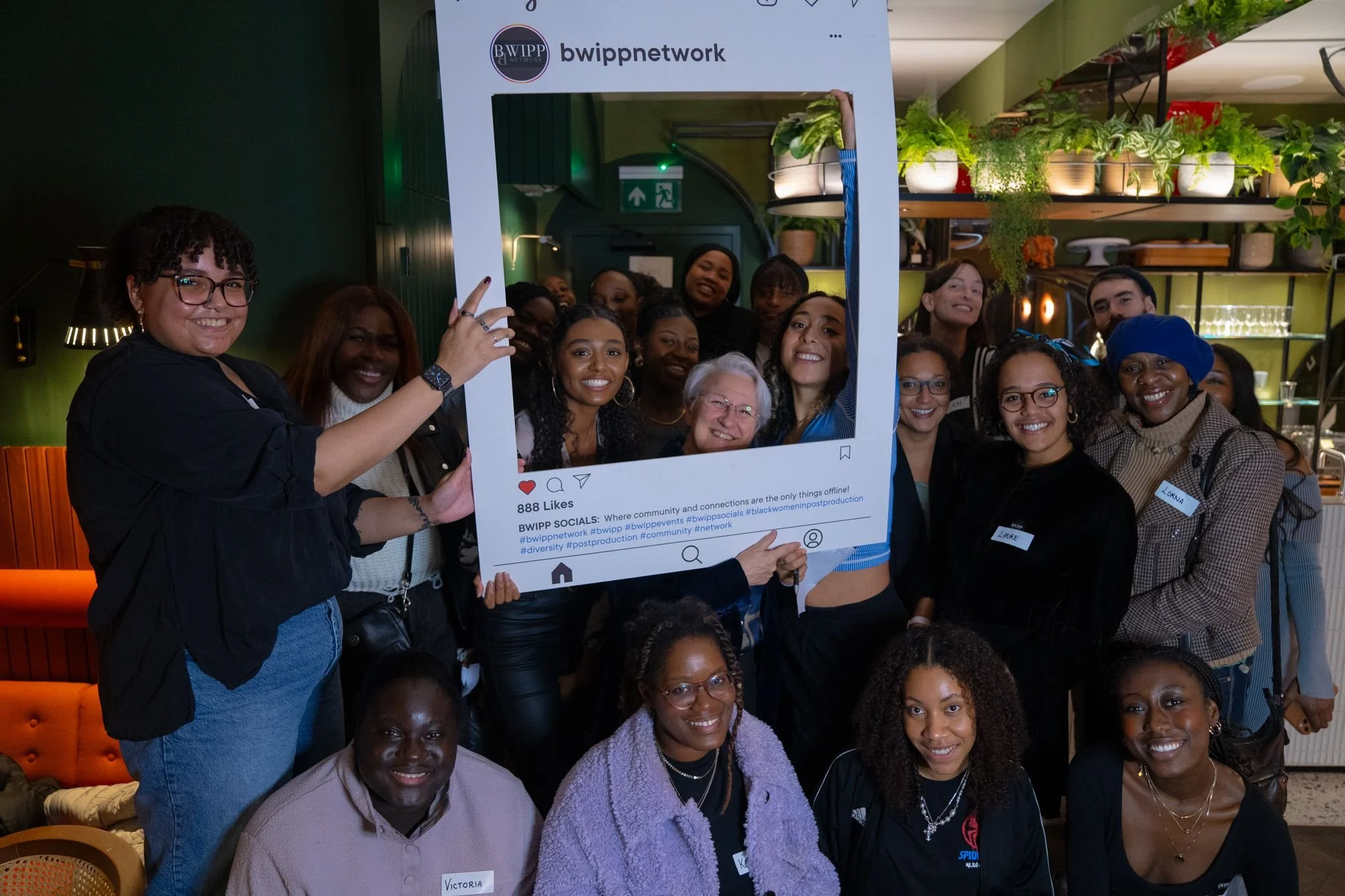 Group of diverse women smiling and posing behind a large social media-style photo frame with the handle @bwippnetwork, in a green-themed restaurant or cafe with shelves of plants and kitchen items in the background.