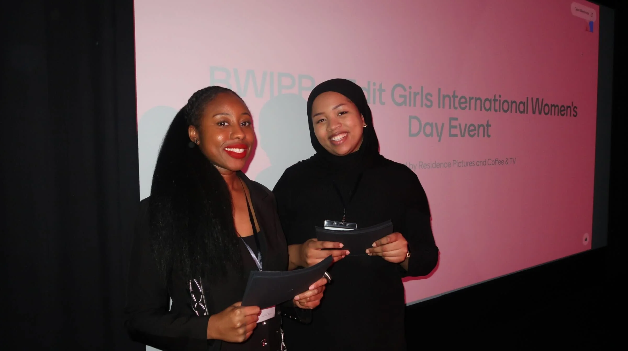Two women smiling and holding papers, standing in front of a pink screen that reads "BWP Girls International Women's Day Event."