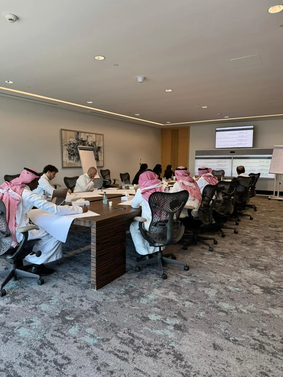 A group of men dressed in traditional Middle Eastern attire and some in white shirts, seated around a conference table in a modern meeting room, watching a presentation on large screens.