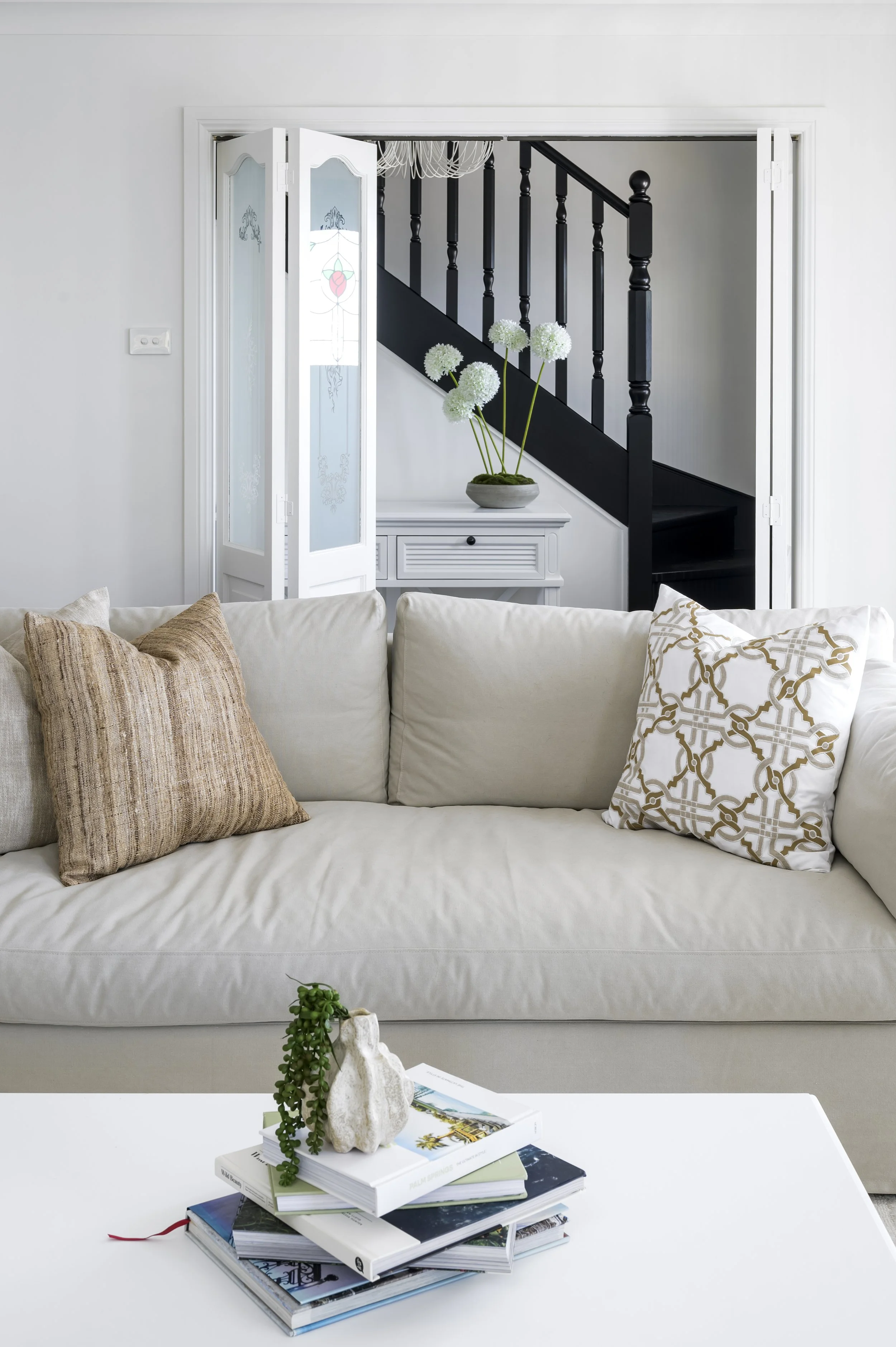 Living room with white sofa, beige and patterned throw pillows, coffee table with books and decorative plant, and an open doorway revealing a staircase with black banister and white walls.