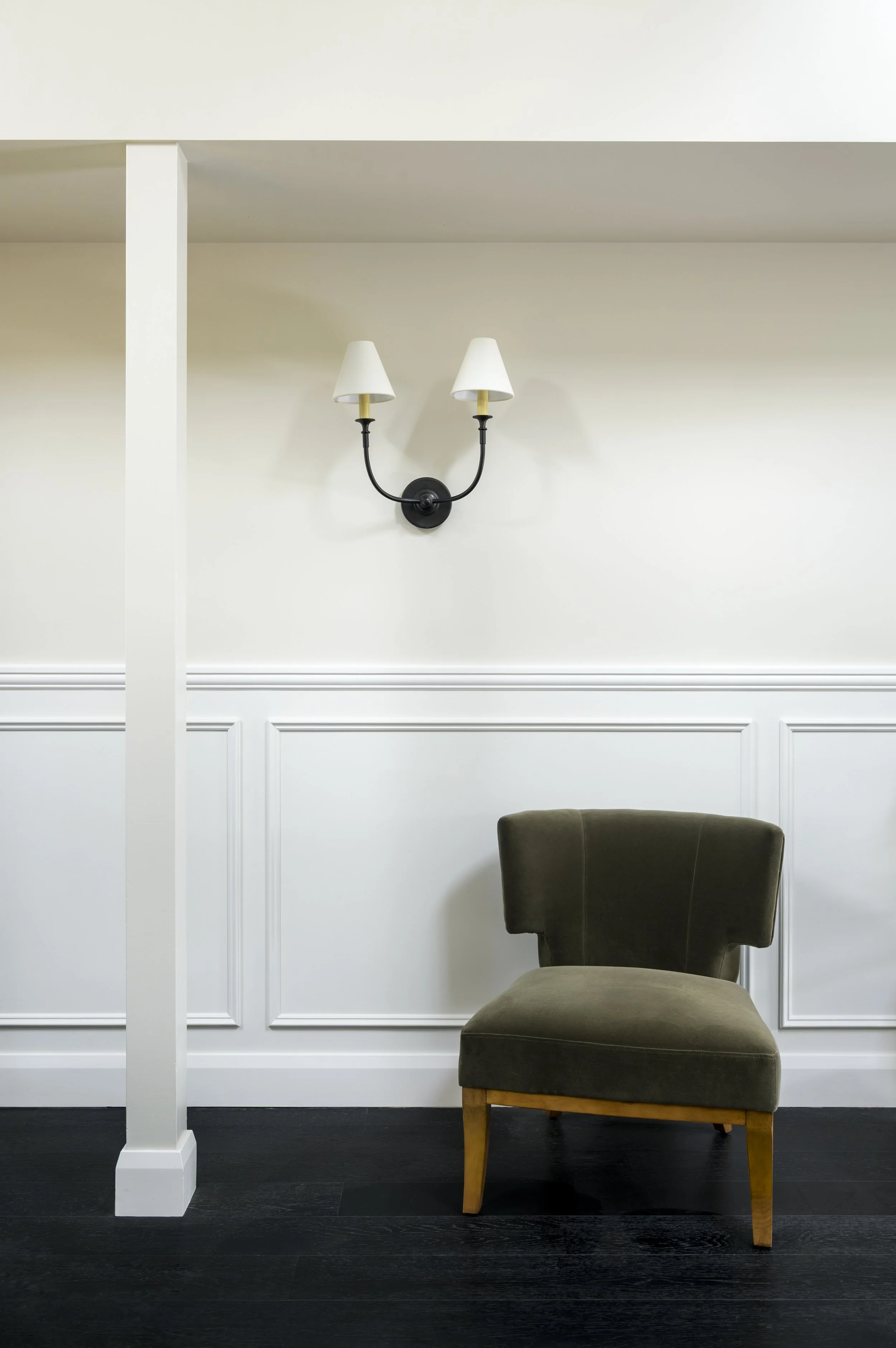 A vintage green upholstered armchair with wooden legs is positioned against a white wall with decorative paneling. Above it, a black wall sconce with two white lampshades is mounted on the wall.