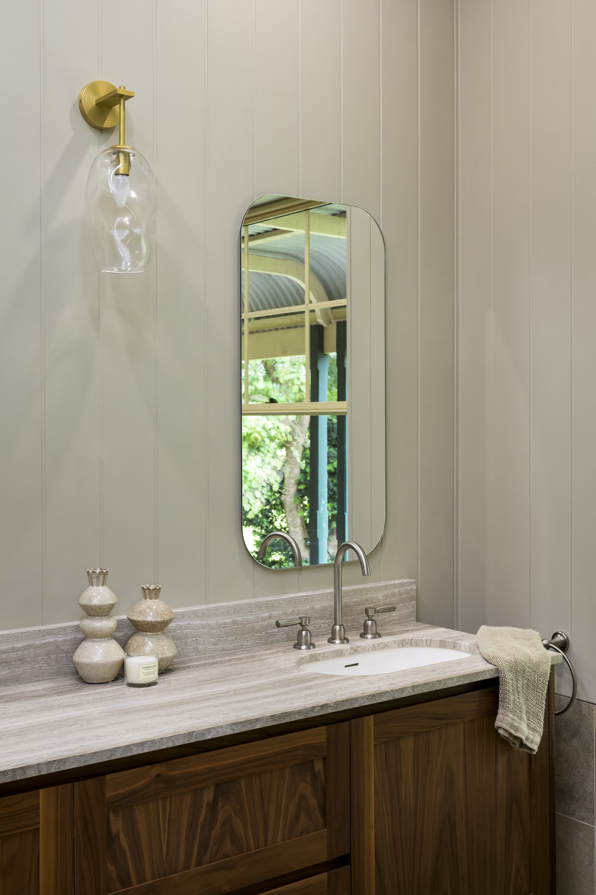Bathroom sink with a mirror, decorative vases, a candle, and a towel, with a window reflecting greenery outside.