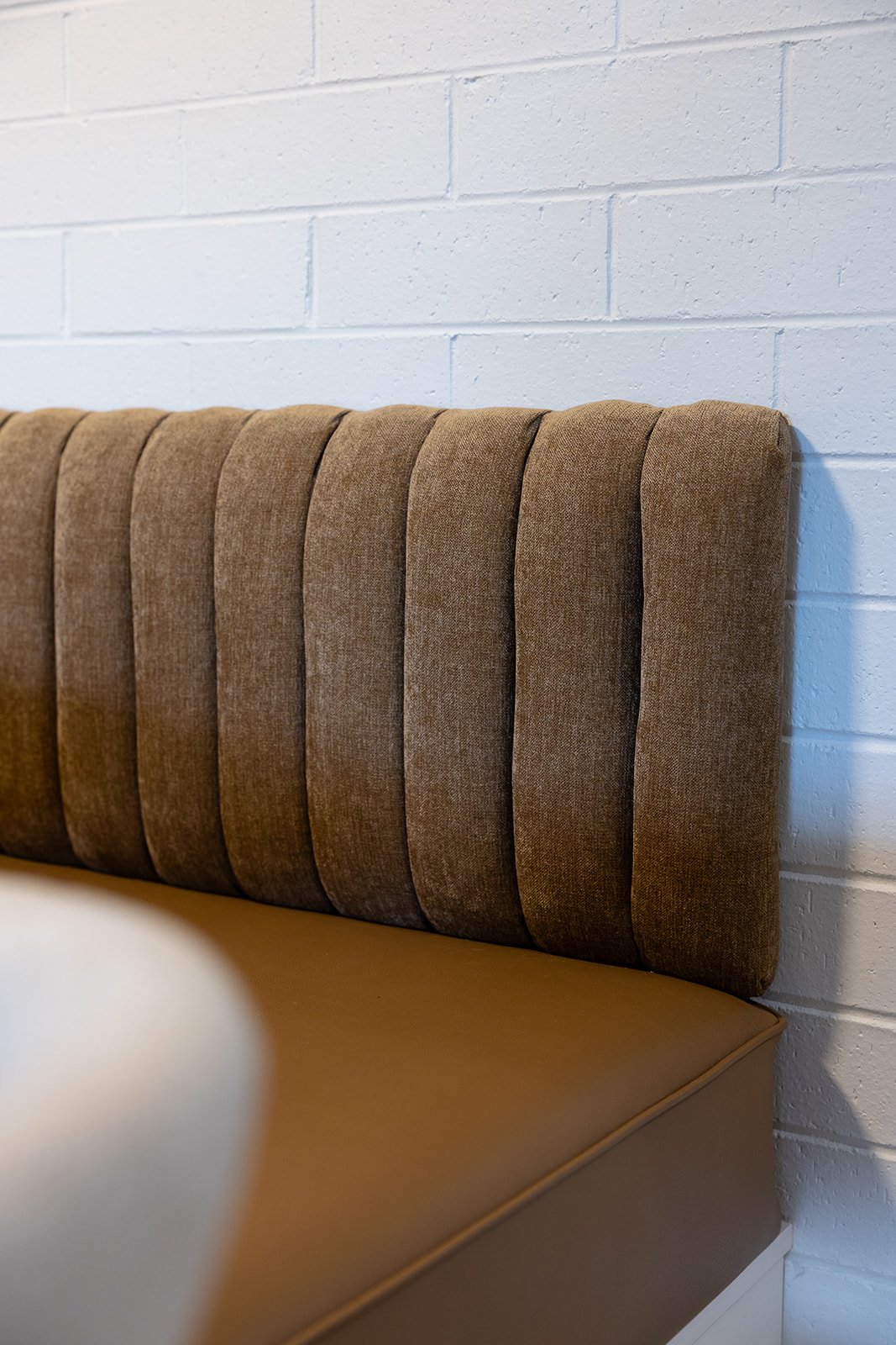 A brown upholstered bench with a high backrest against a white brick wall.