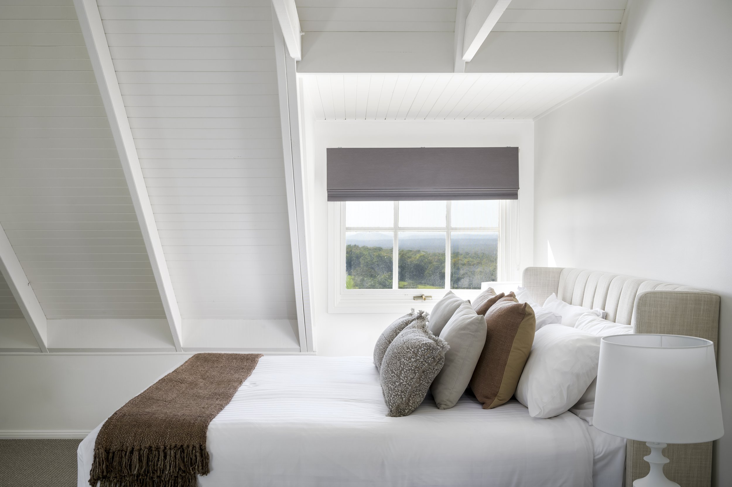 A cozy bedroom with a large bed covered in white linens and multiple decorative pillows. A brown throw blanket is at the foot of the bed. A white nightstand with a white lamp is beside the bed. A window with a view of trees and hills is behind the bed, with a grey shade partially drawn. The room has white walls and a sloped ceiling.