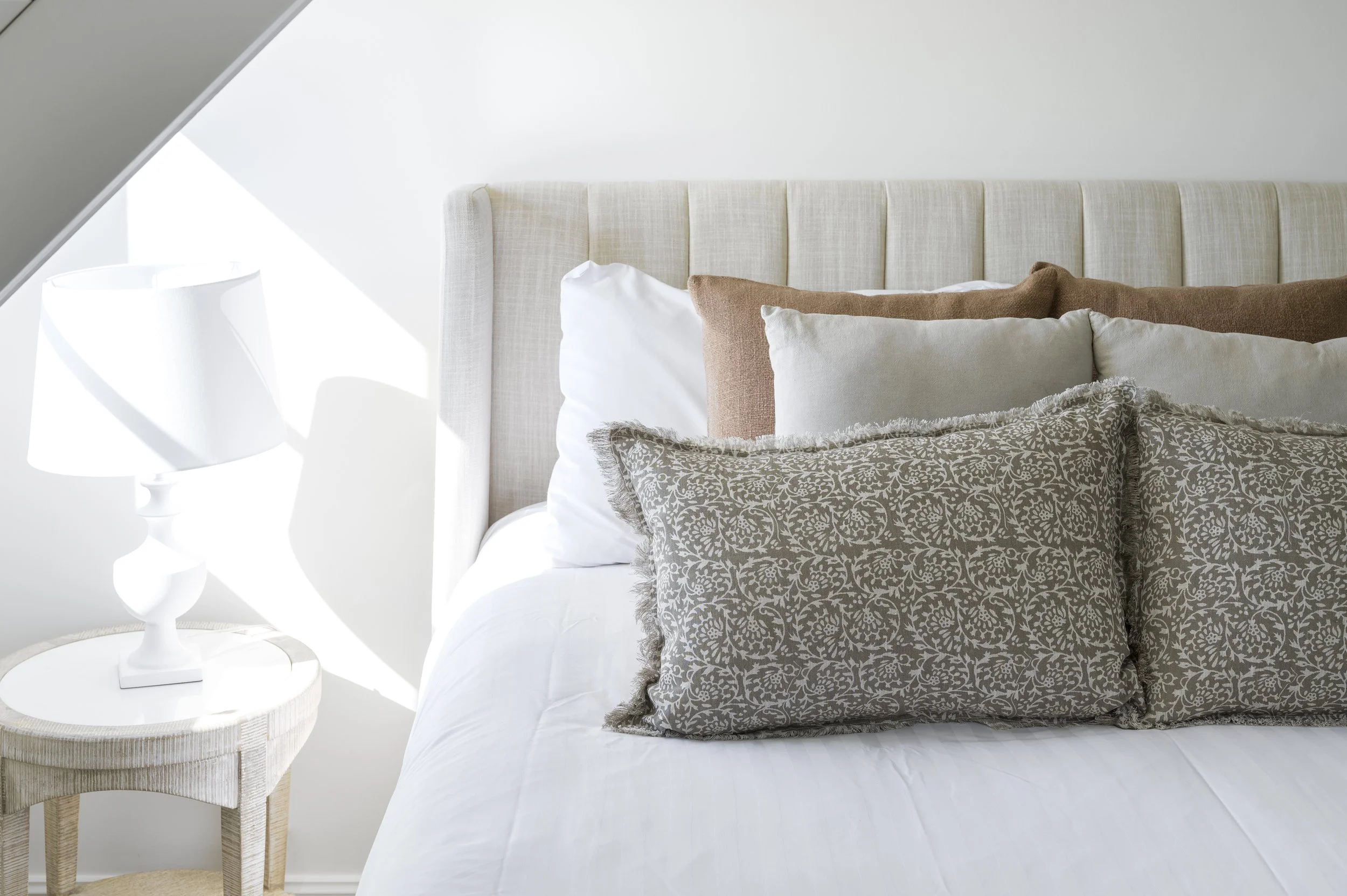 A neatly made bed with multiple decorative pillows, next to a light-colored bedside table with a modern white lamp, in a bright bedroom with natural sunlight.