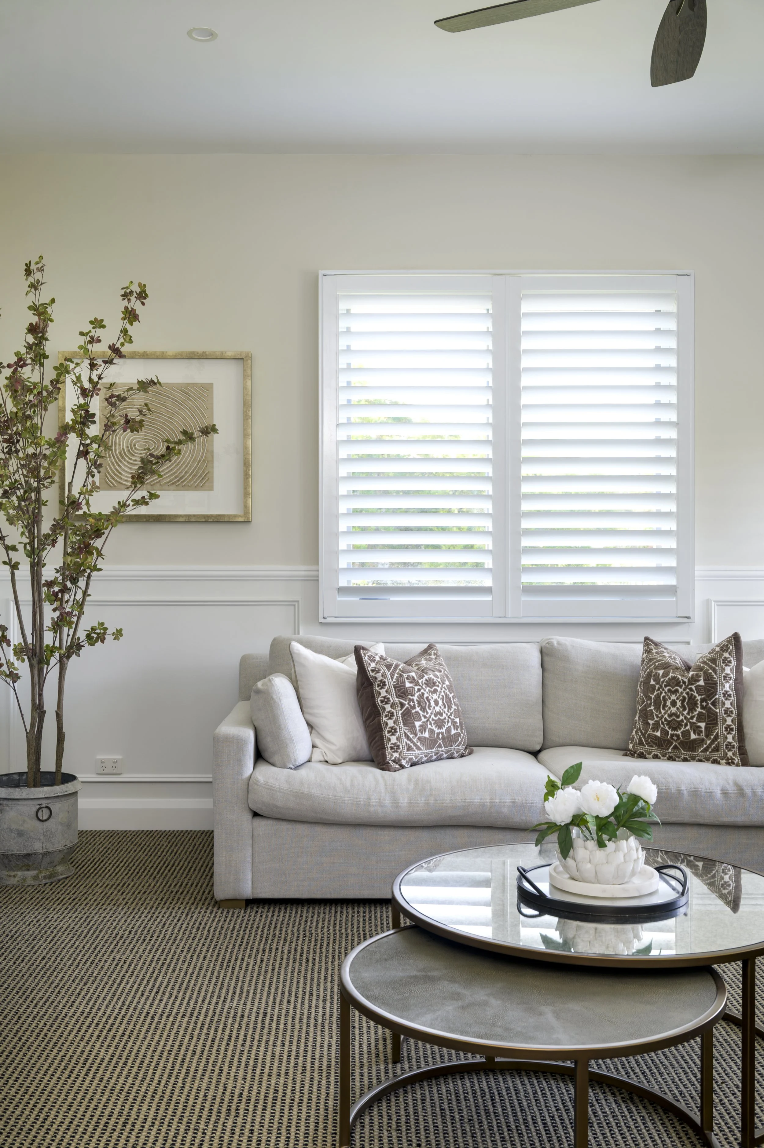 Bright living room with large window, white shutters, beige sofa with throw pillows, potted plant, framed artwork, and coffee table with flower arrangement.