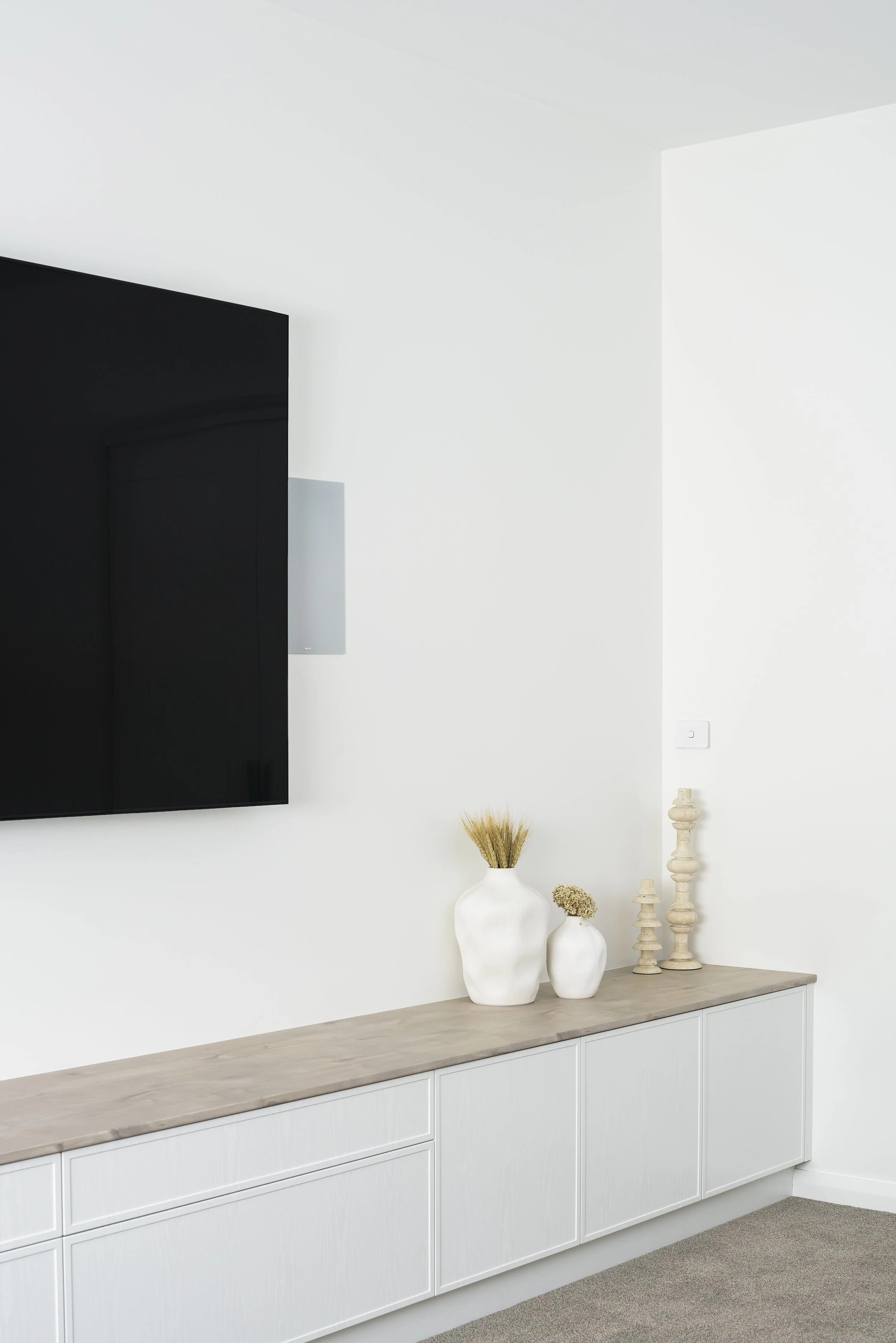A minimalist interior with a wall-mounted TV, white sideboard, and decorative vases and wooden sculptures.