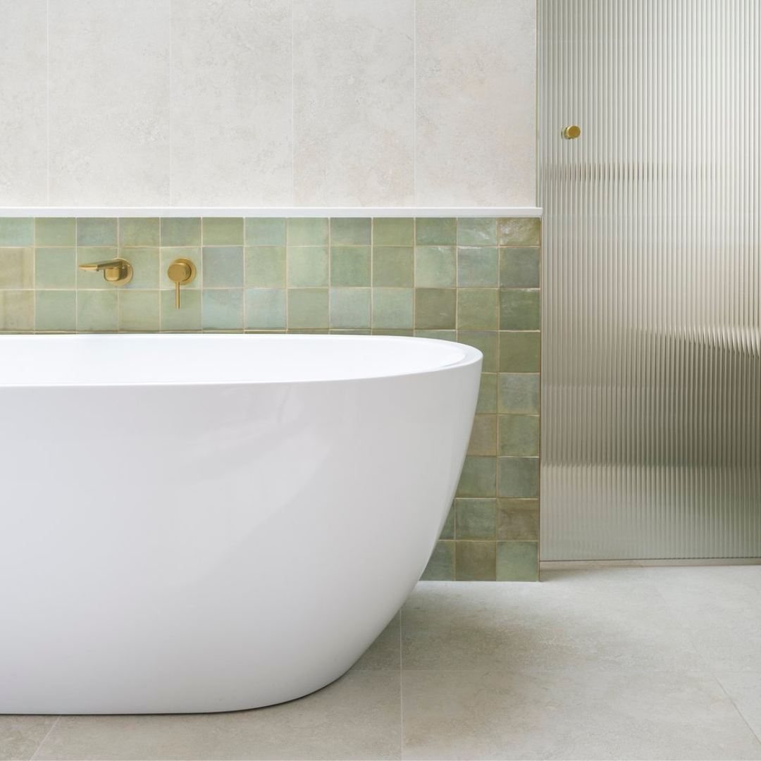 Modern bathroom featuring a white freestanding bathtub, gold fixtures, green tiled accent wall, marble wall tiles, and a textured frosted glass door.