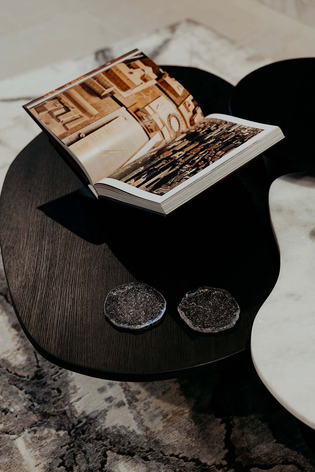 An open magazine on a black coffee table, with two glass paperweights nearby, against a backdrop of a light-colored rug and marble flooring.