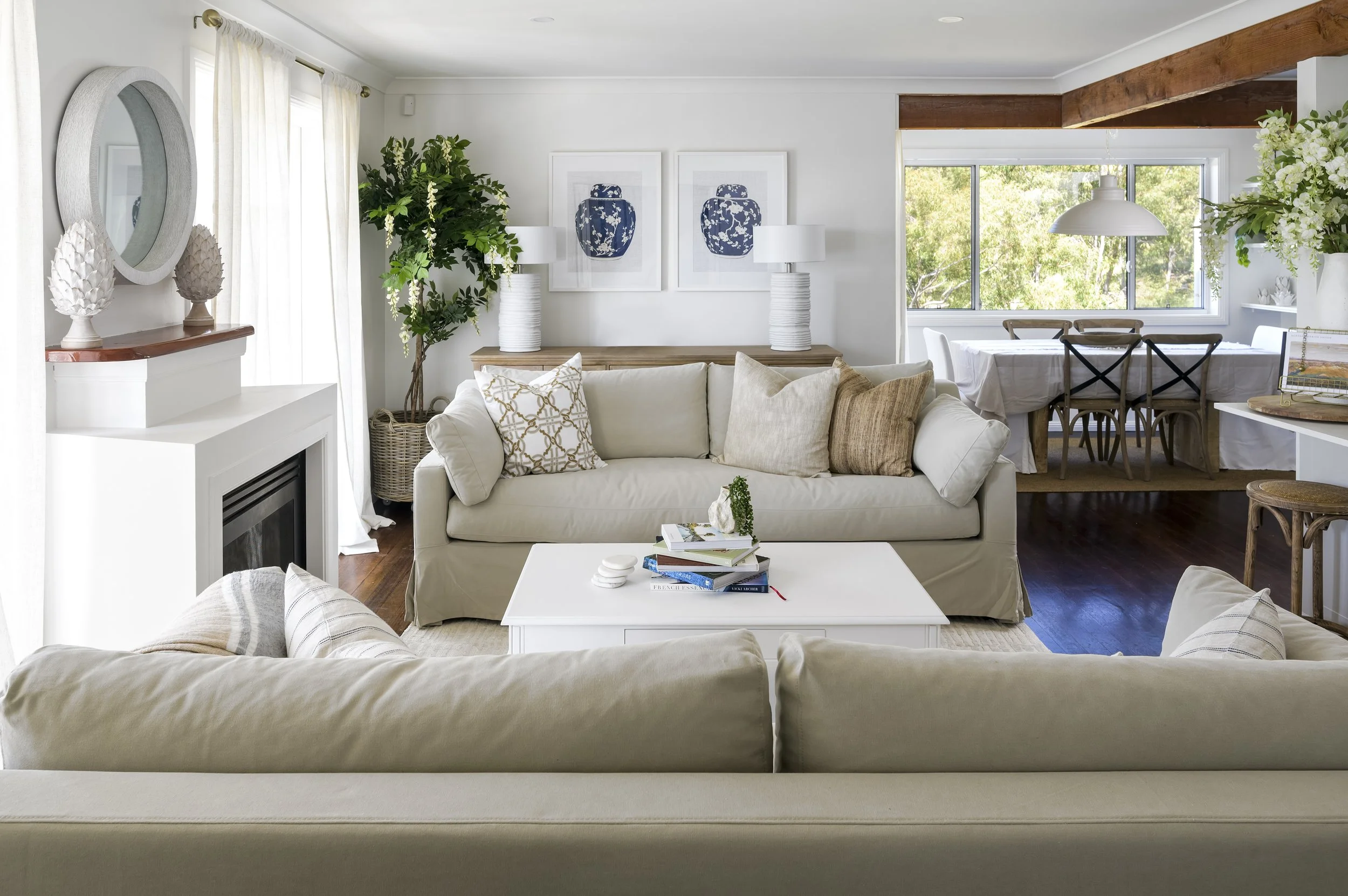 Bright living room with white walls, beige sofas, a white coffee table with books, and a dining area with a large window and wooden chairs in the background.