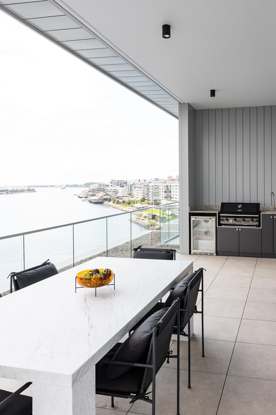 Modern outdoor balcony with a white marble dining table, black chairs, a fruit bowl, built-in grill, mini fridge, and glass railing overlooking city and water.