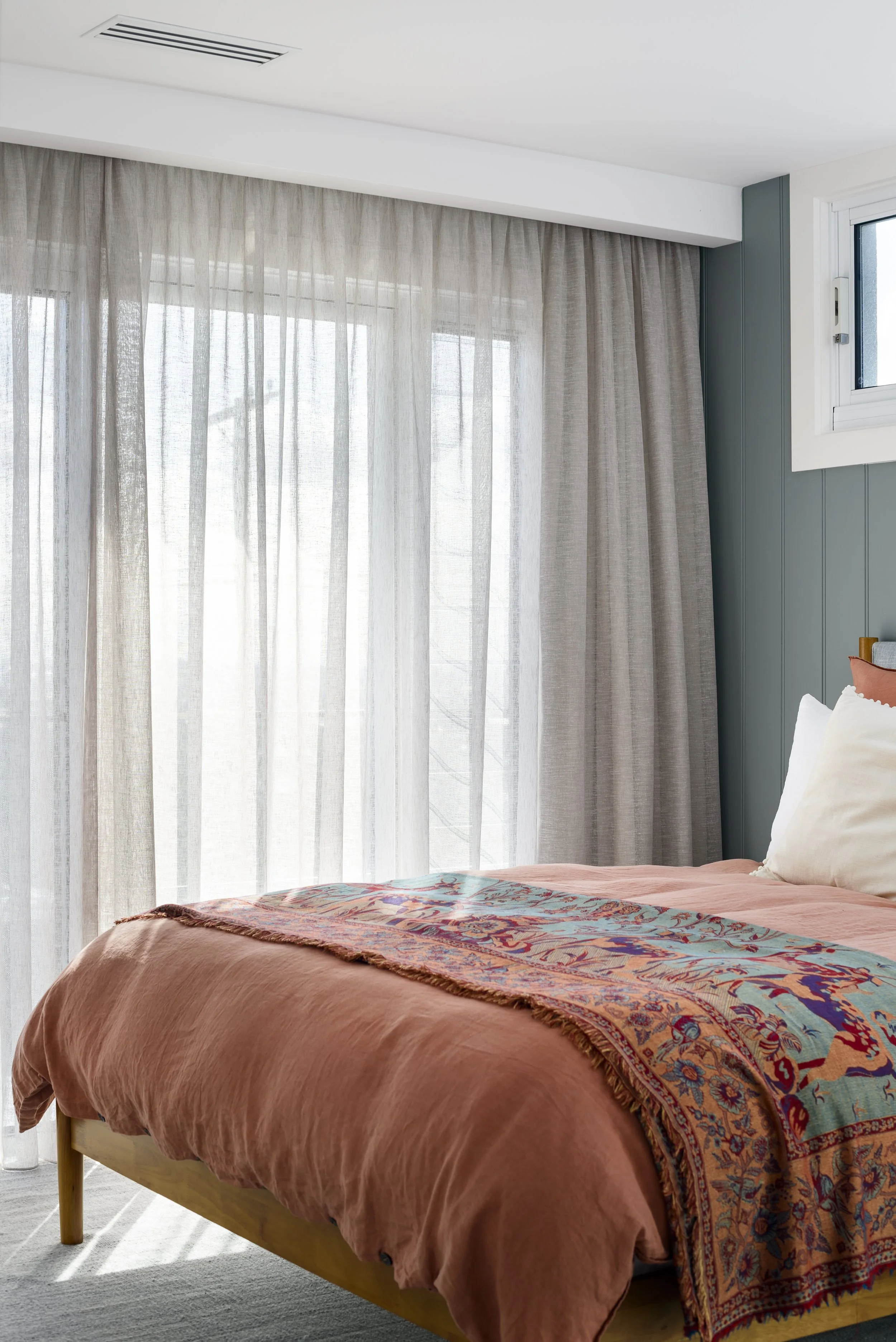 A bedroom with a bed dressed in peach-colored bedding and a colorful throw blanket. Large windows with sheer curtains allow natural light into the room. A small window is visible on the right.