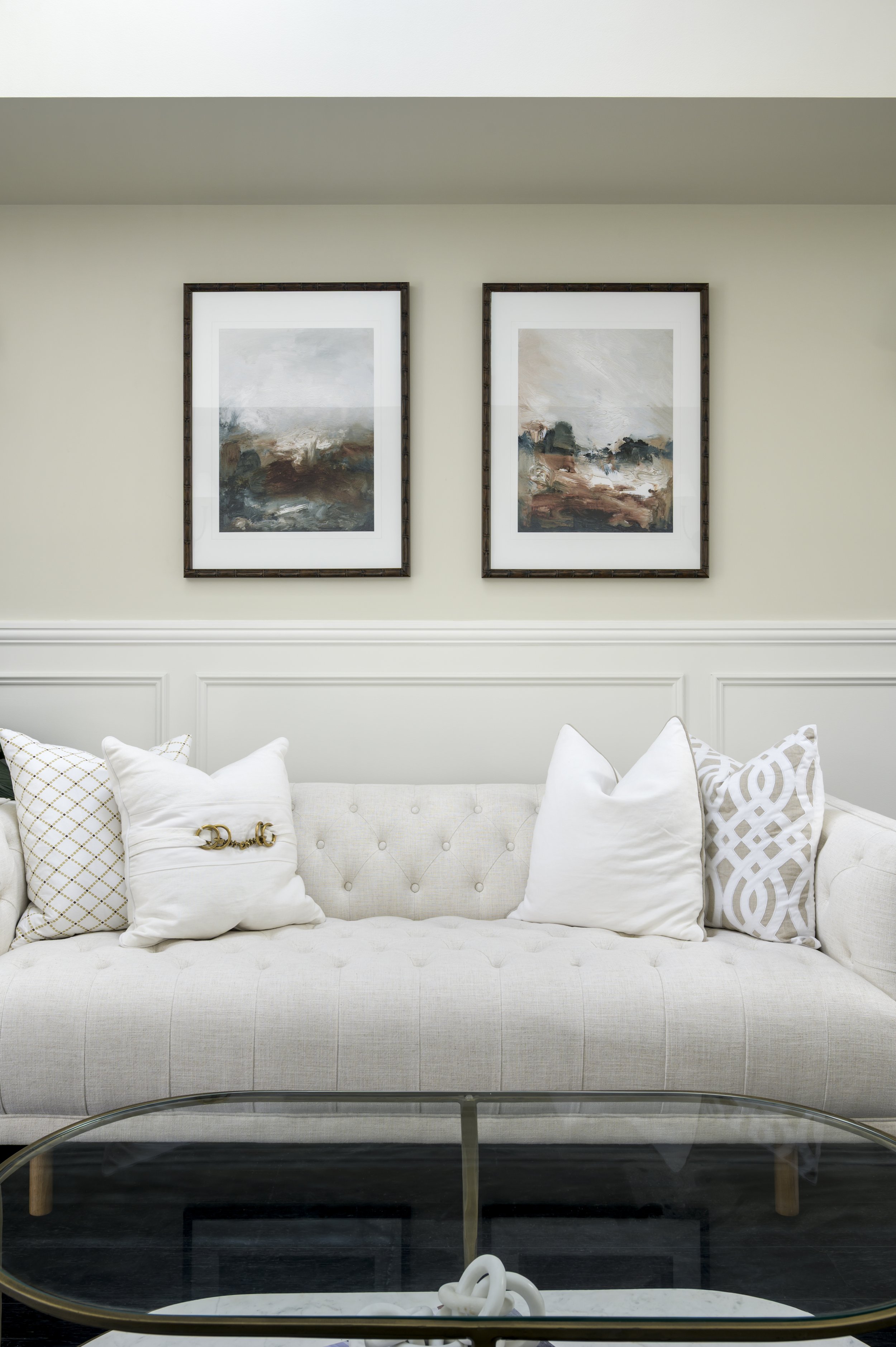 A cream-colored tufted sofa with white patterned and plain pillows, situated in front of a wall with two framed abstract landscape paintings, and a glass coffee table with a white decorative ornament.
