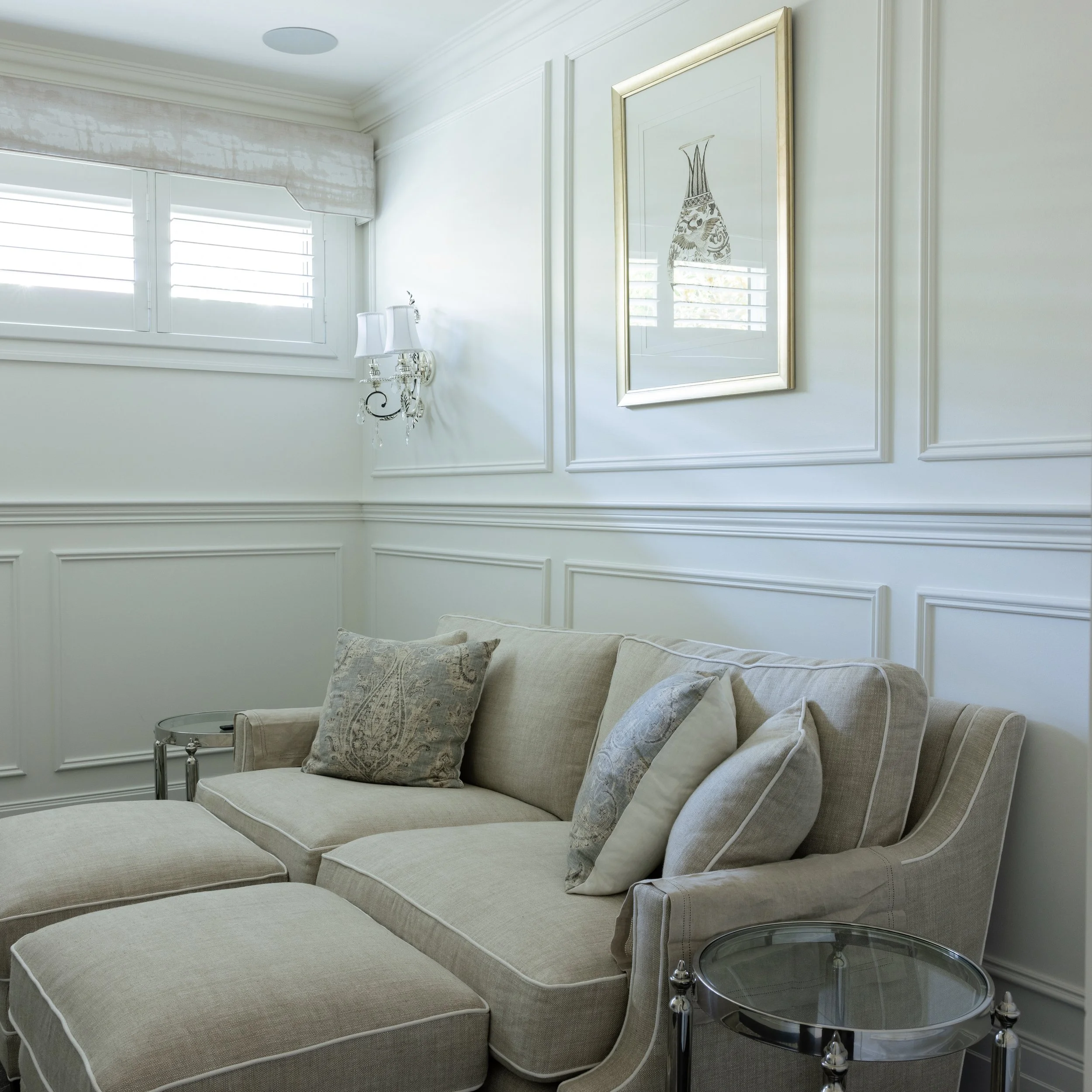 A beige sectional sofa with patterned pillows next to glass side tables in a bright living room with white panel walls, a window with shutters, and a framed art piece on the wall.