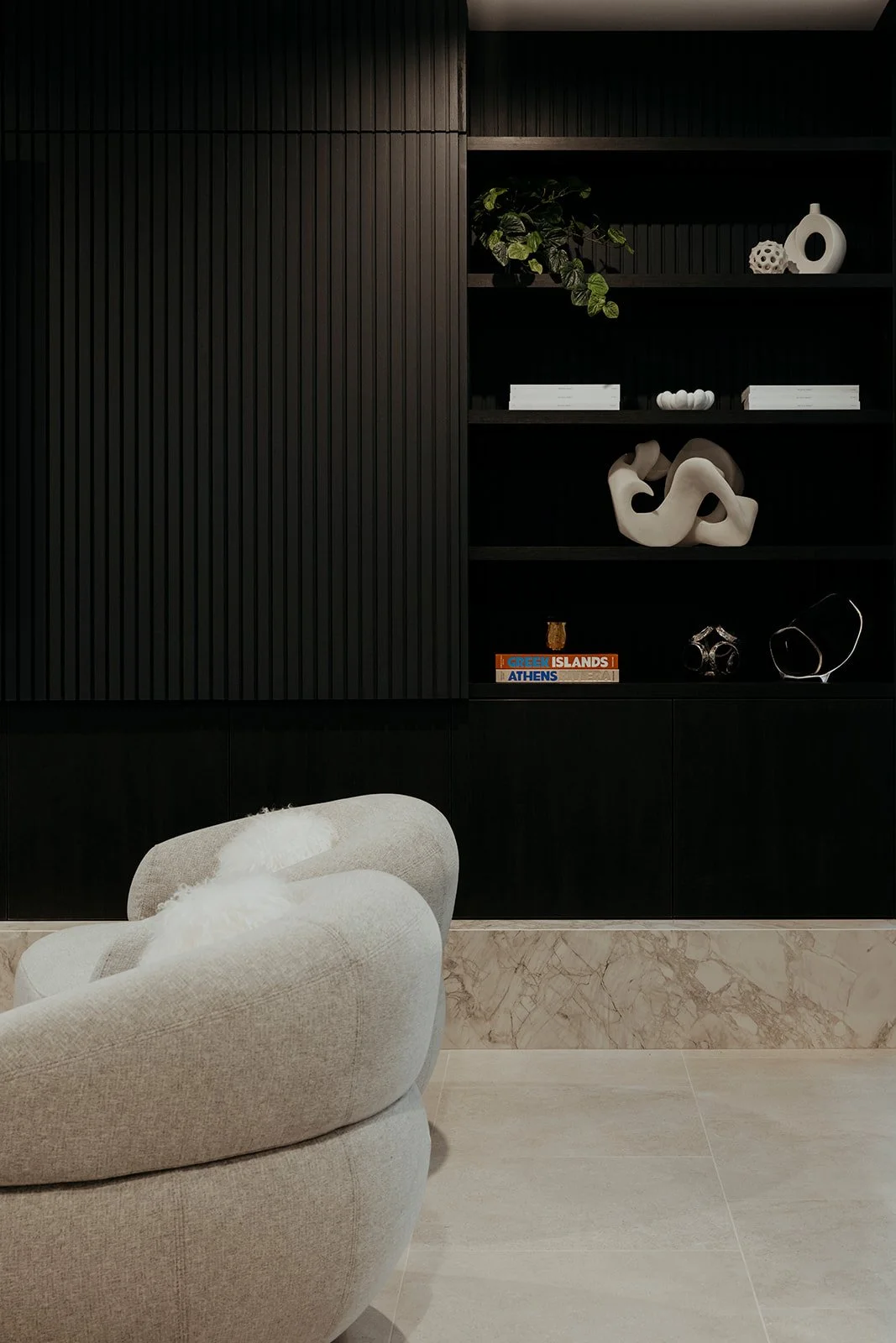 Modern living room with a beige upholstered armchair in front of black wooden shelving filled with decorative objects and books, and a light-colored marble wall and floor.