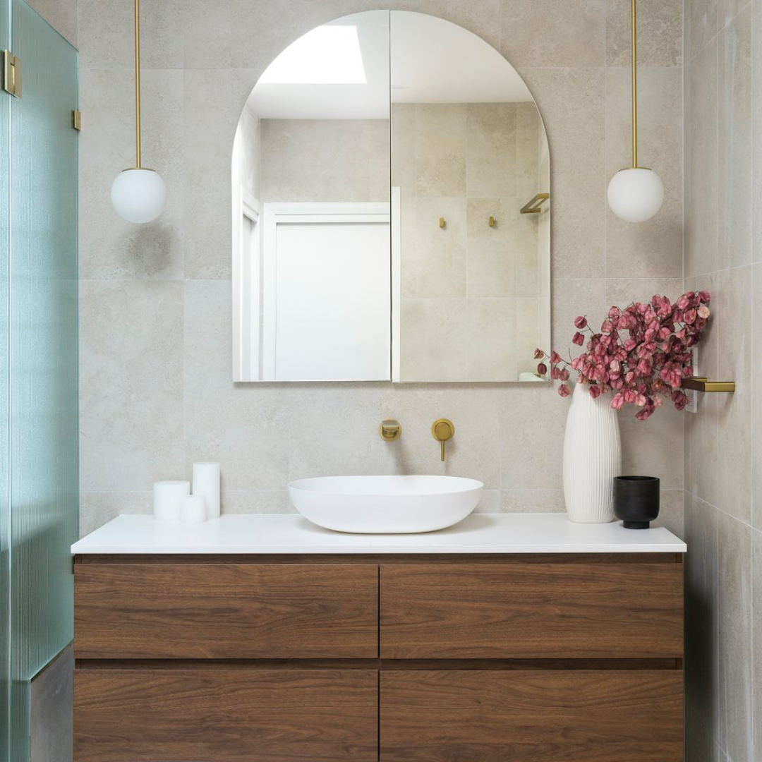 Modern bathroom with a wooden vanity, oval vessel sink, large mirror, wall-mounted faucet, pink floral arrangement in a white vase, black container, and two hanging globe pendant lights.