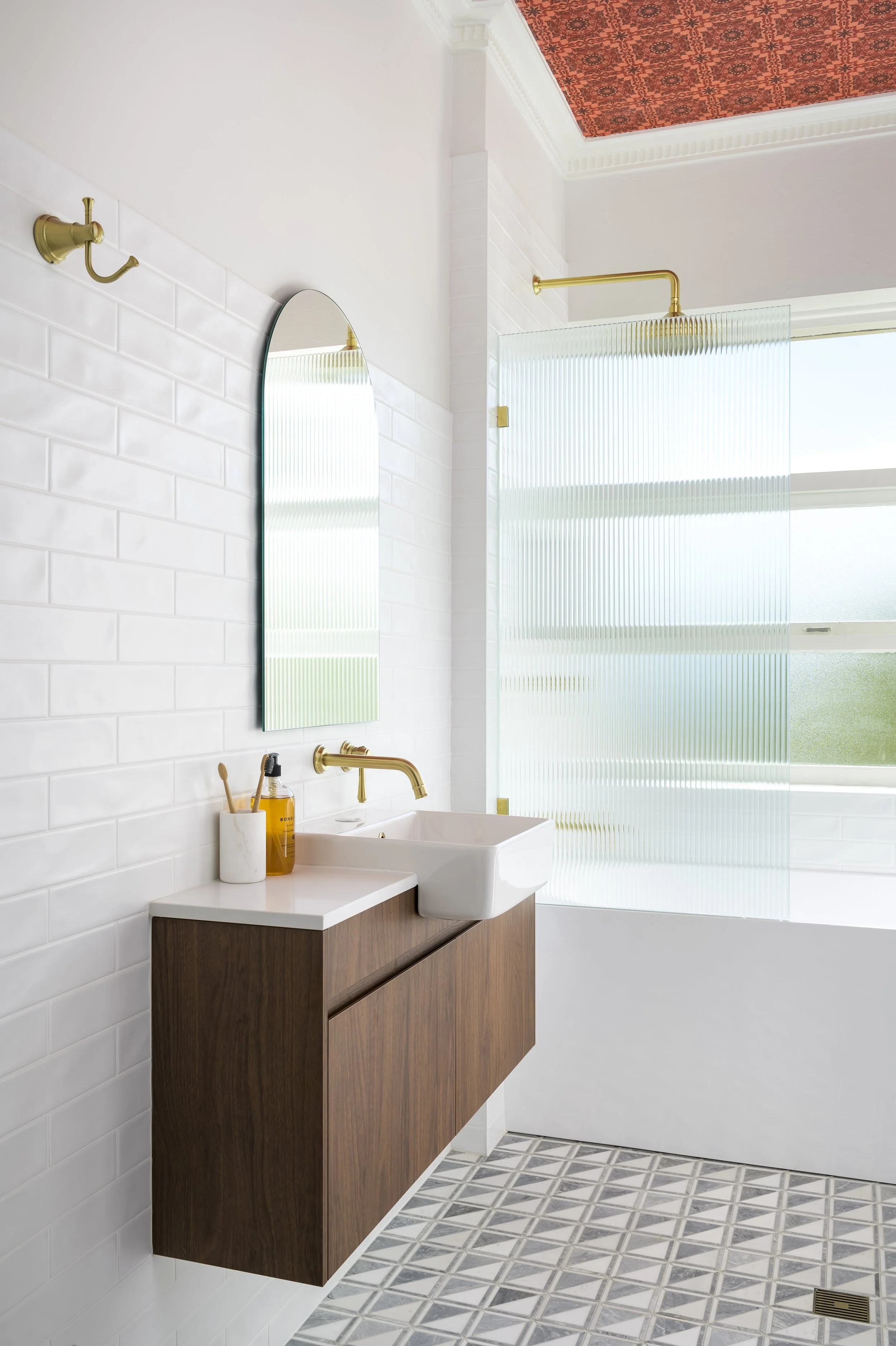 Modern bathroom with white tiled wall, wooden vanity with a vessel sink, gold fixtures, a tall mirror, patterned floor tiles, and a glass shower enclosure with a frosted window.